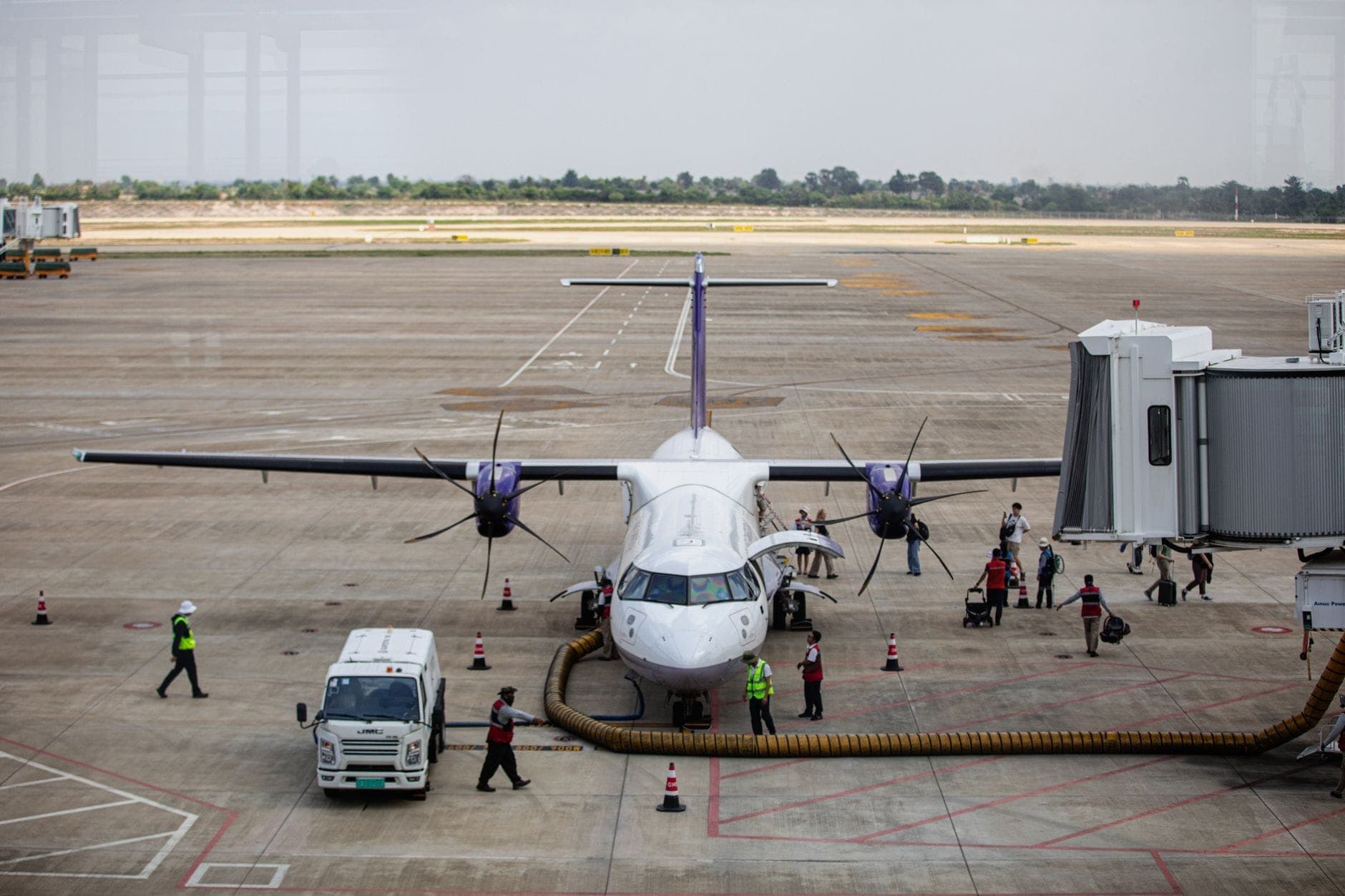 Vista desde el suelo de una pequeña aeronave comercial conectada a una manga de embarque en un aeropuerto en Tailandia, con personal y pasajeros embarcando y desembarcando.