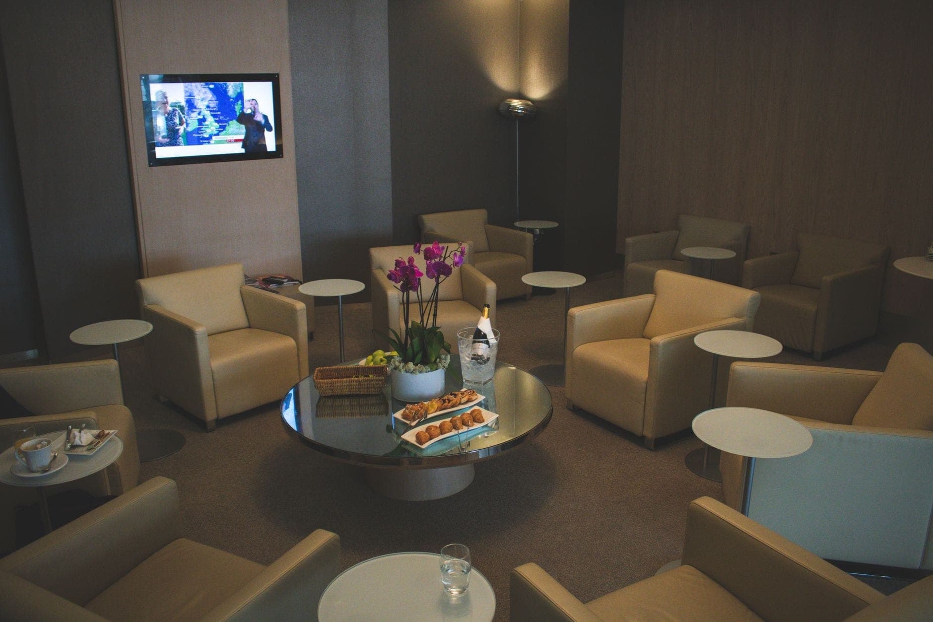 Salón moderno de aeropuerto con sillones beige, mesas pequeñas, un televisor y refrigerios sobre una mesa central de cristal.