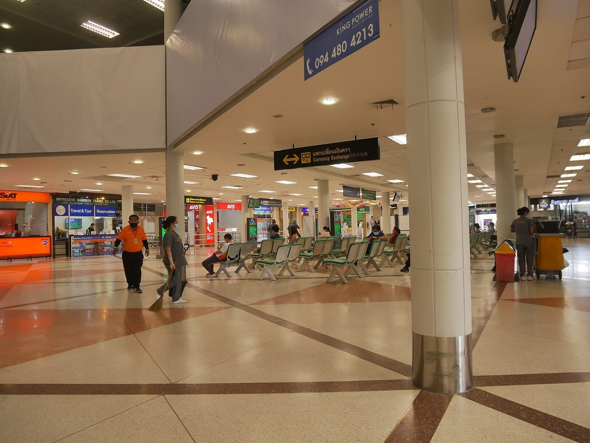 Área de asientos en el Aeropuerto Internacional de Chiang Mai con viajeros, personal de limpieza y letreros de alquiler de autos, cambio de divisas y reservas de tours.