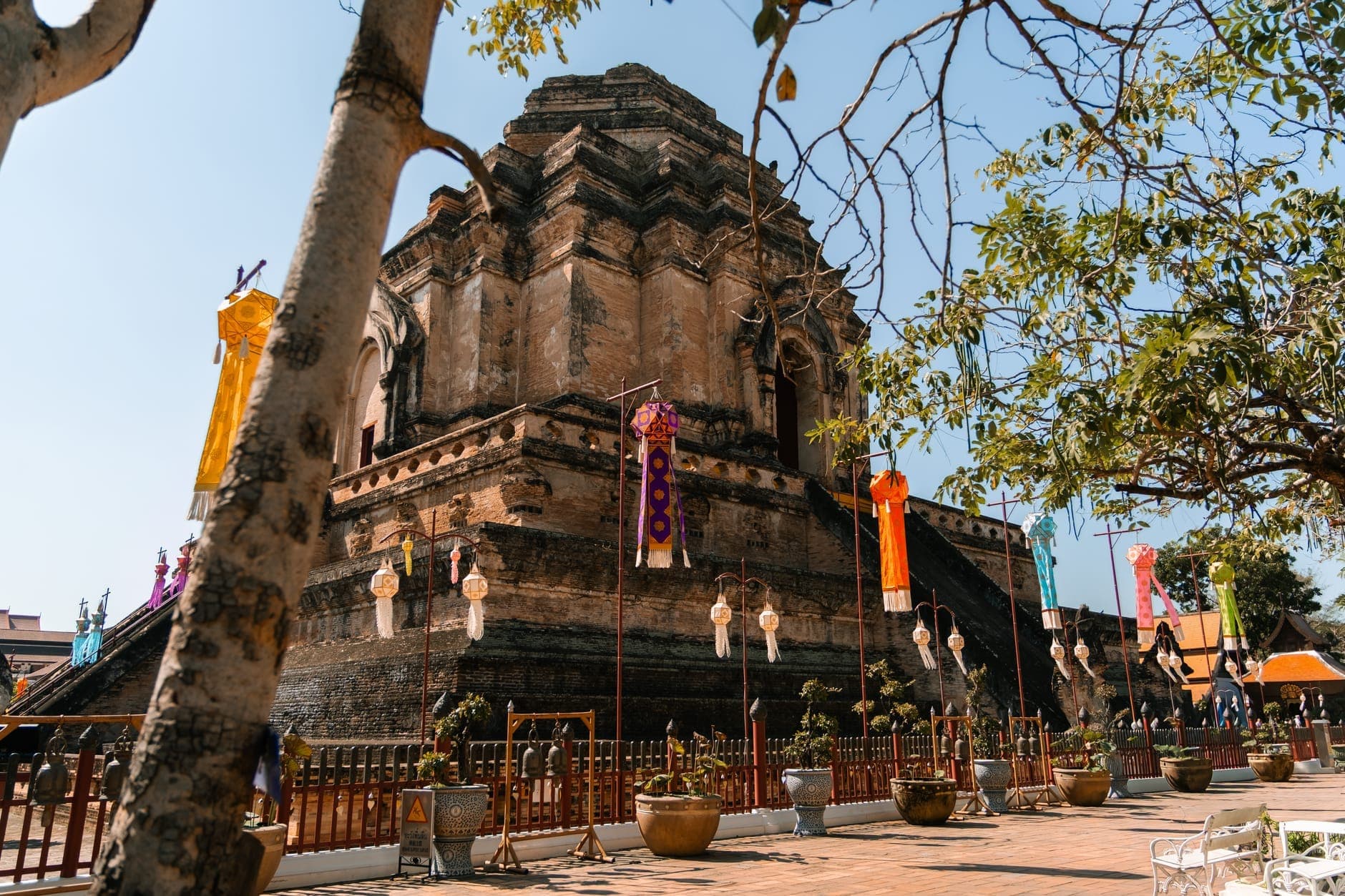 Ancient brick stupa of Wat Chedi Luang in Chiang Mai, surrounded by colorful lanterns and trees under a clear blue sky.