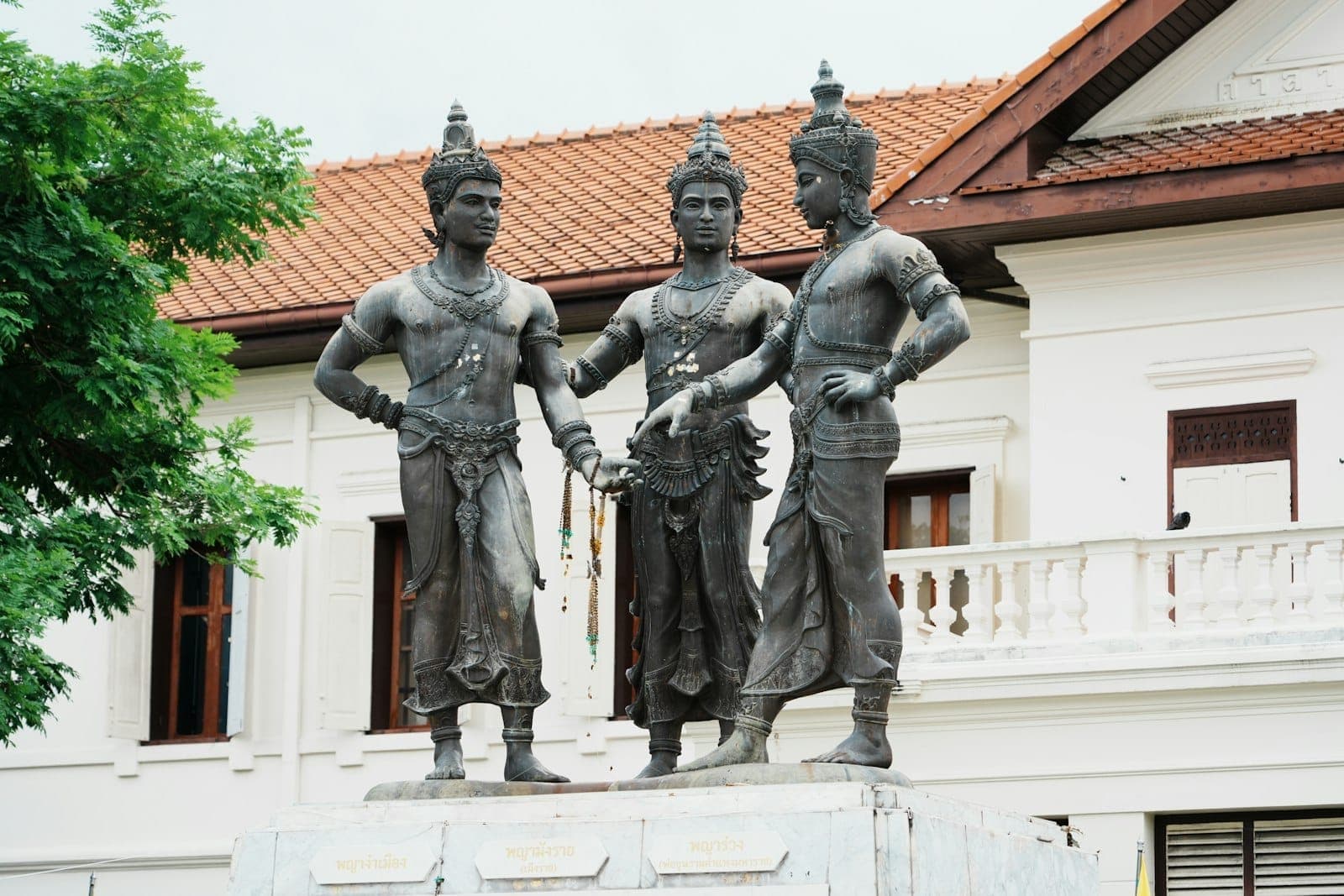 Statue der drei Könige in traditioneller Kleidung vor einem weißen Kolonialgebäude im historischen Zentrum von Chiang Mai.