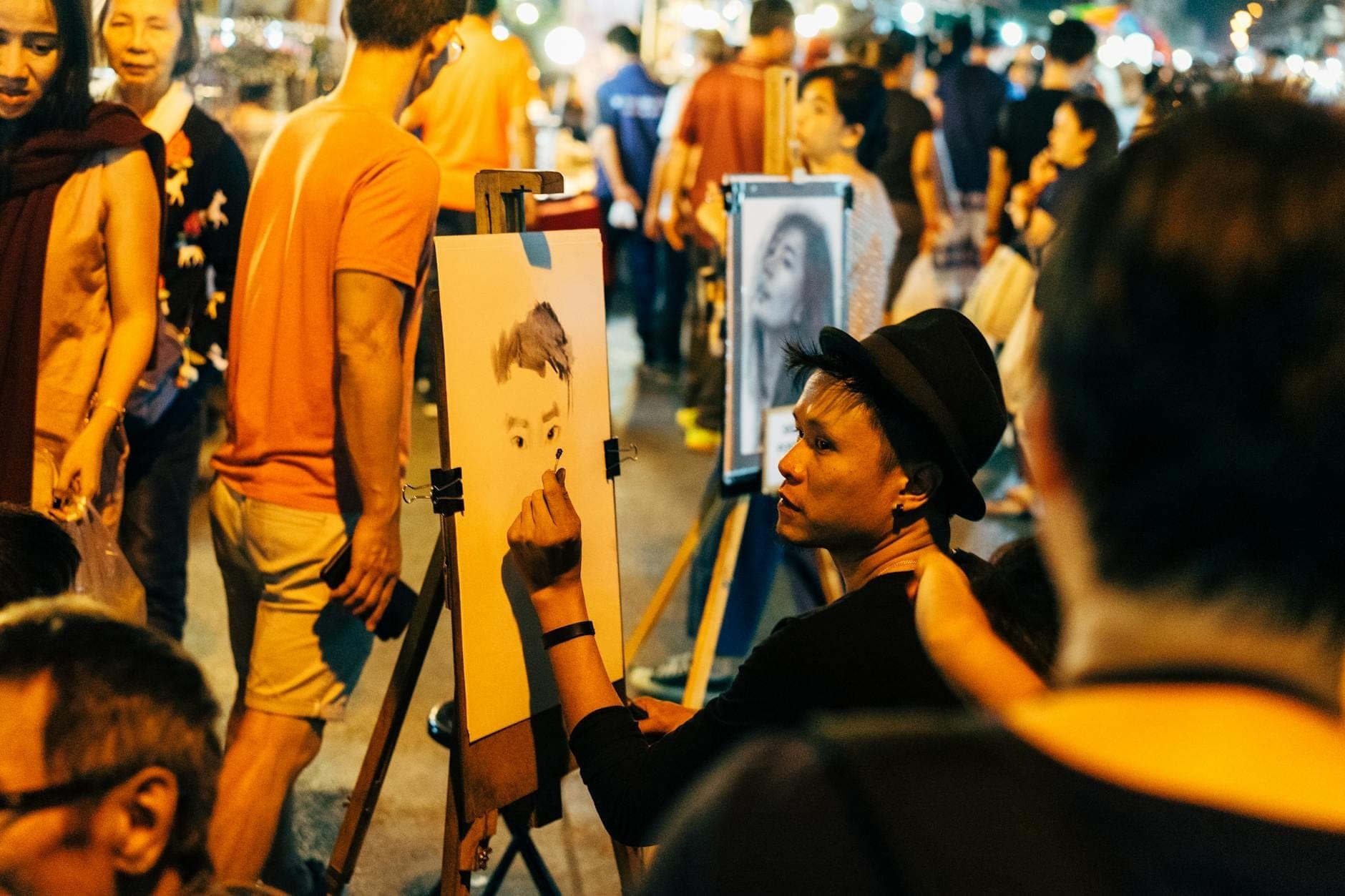 Un artista di strada che disegna un ritratto dal vivo all'aperto di notte, circondato da persone e dalle vivaci luci del mercato.