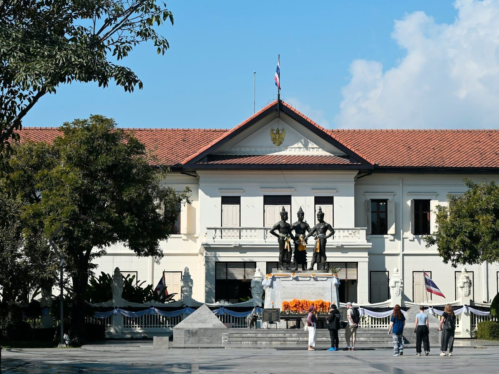 Kolonialstil-Fassade des Chiang Mai City Arts and Cultural Centre mit dem Drei-Könige-Monument, umgeben von Bäumen und Besuchern an einem sonnigen Tag.