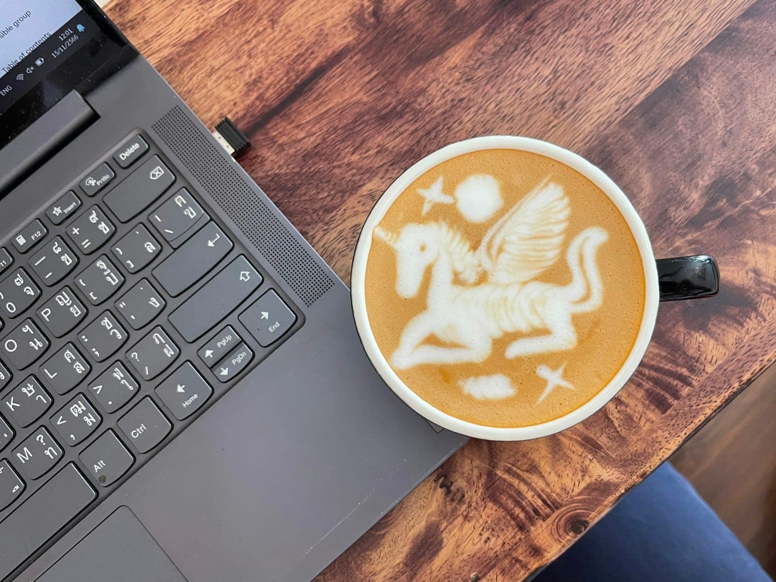 Latte z motywem jednorożca obok laptopa na drewnianym stole, sugerujące przytulne miejsce pracy w kawiarni.