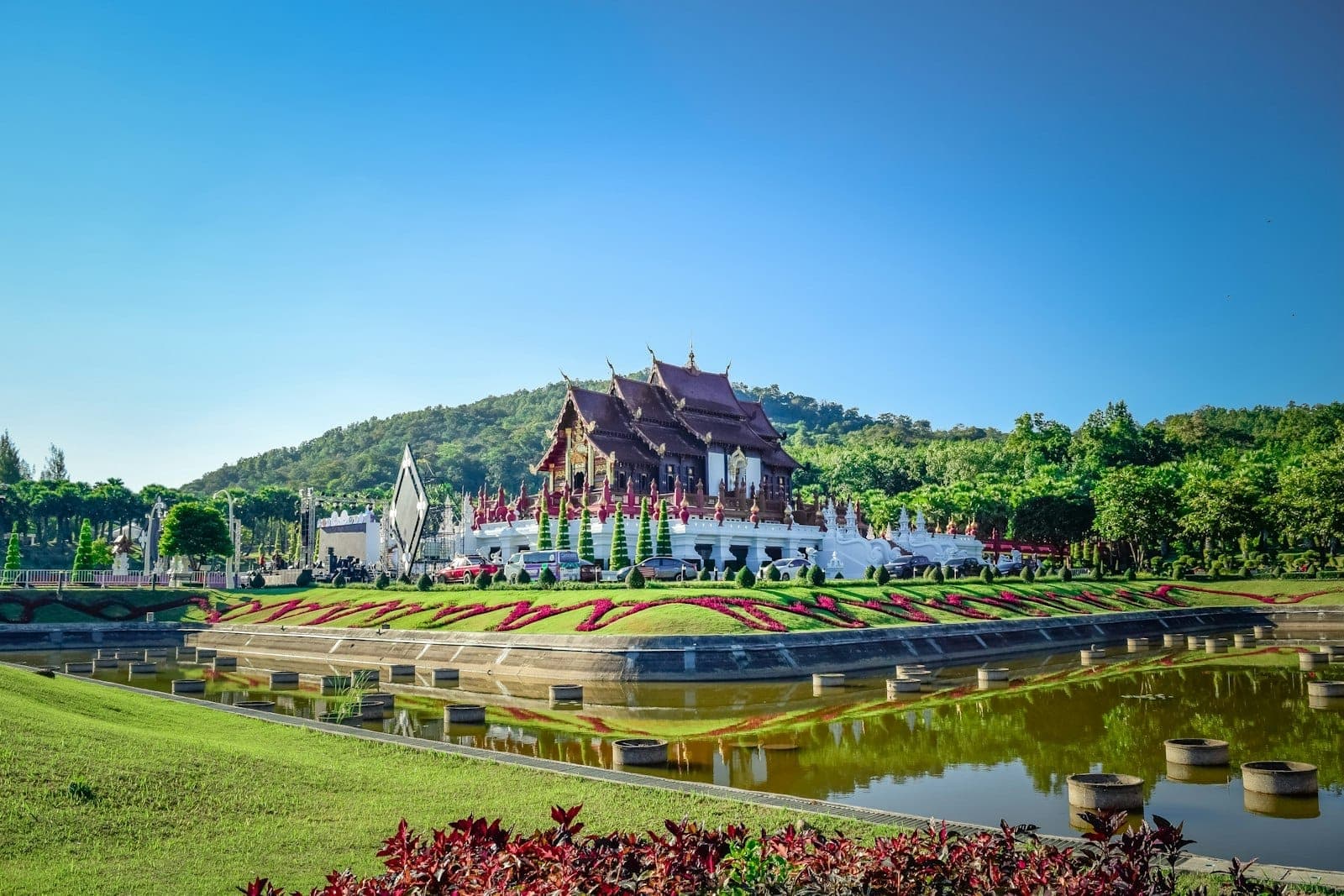 Pabellón de templo de estilo Lanna ornamentado rodeado de jardines y estanque reflectante en el Royal Park Rajapruek, Chiang Mai, bajo un cielo azul despejado.
