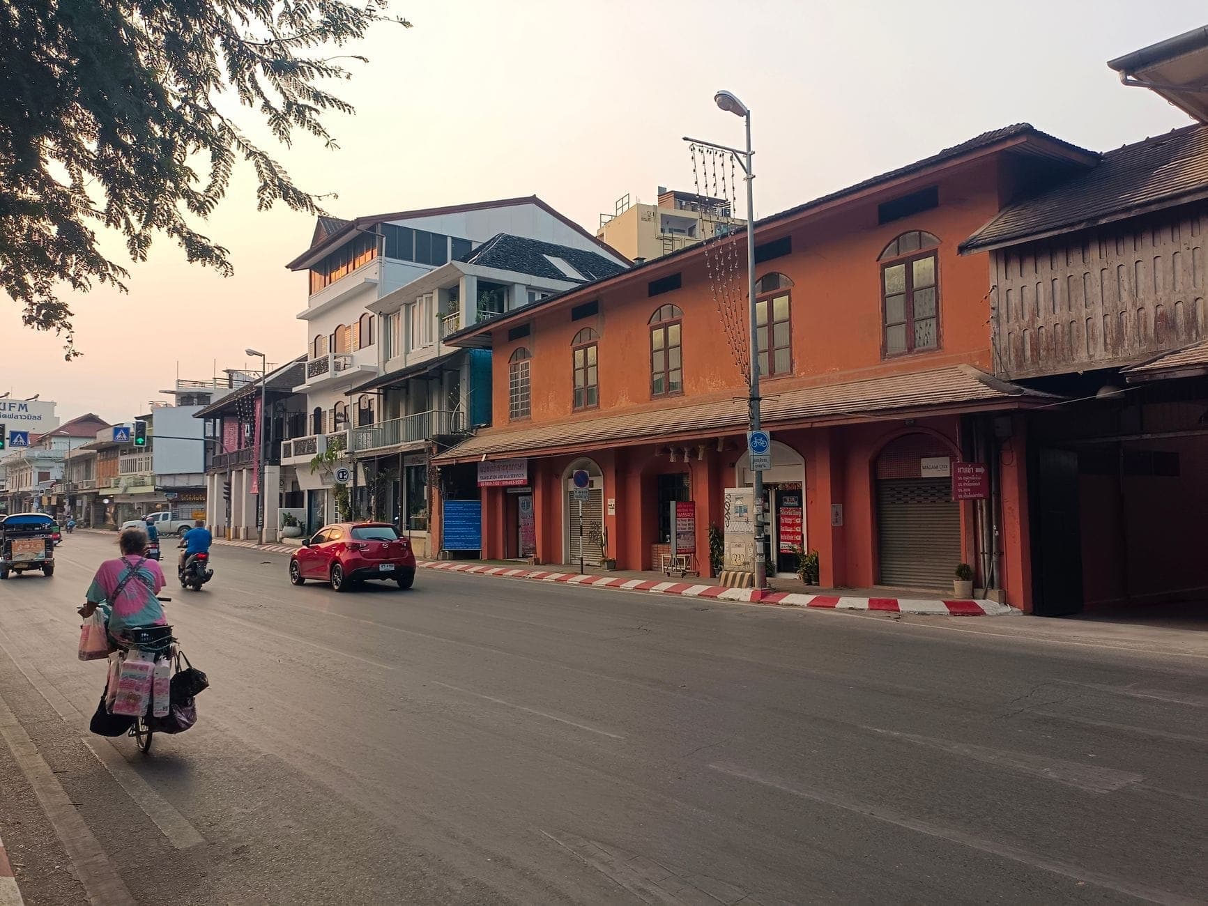 Eine ruhige Straße in Chiang Mai bei Sonnenaufgang mit bunten Gebäuden, einem Radfahrer und lokalem Verkehr, die das entspannte Stadtflair zeigt.