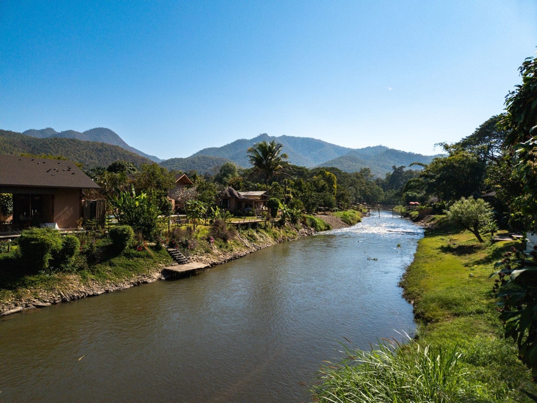 Ein Fluss schlängelt sich durch ein grünes Bergtal mit kleinen Häusern und üppiger Vegetation unter einem strahlend blauen Himmel in der Nähe von Chiang Mai.