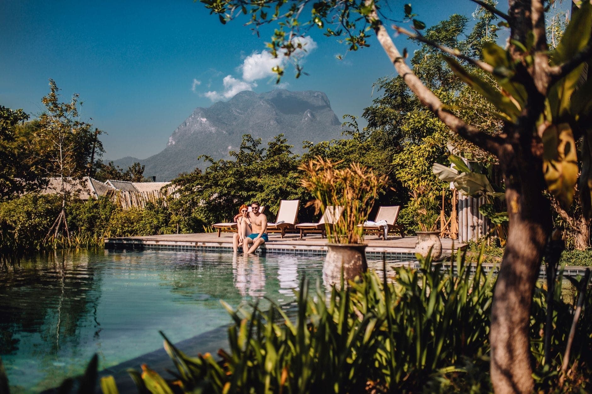 Ein Paar entspannt am Rand eines ruhigen Infinity-Pools mit üppigem Grün und Bergblick in einem Luxusresort in Chiang Mai, Sonnenliegen in der Nähe.