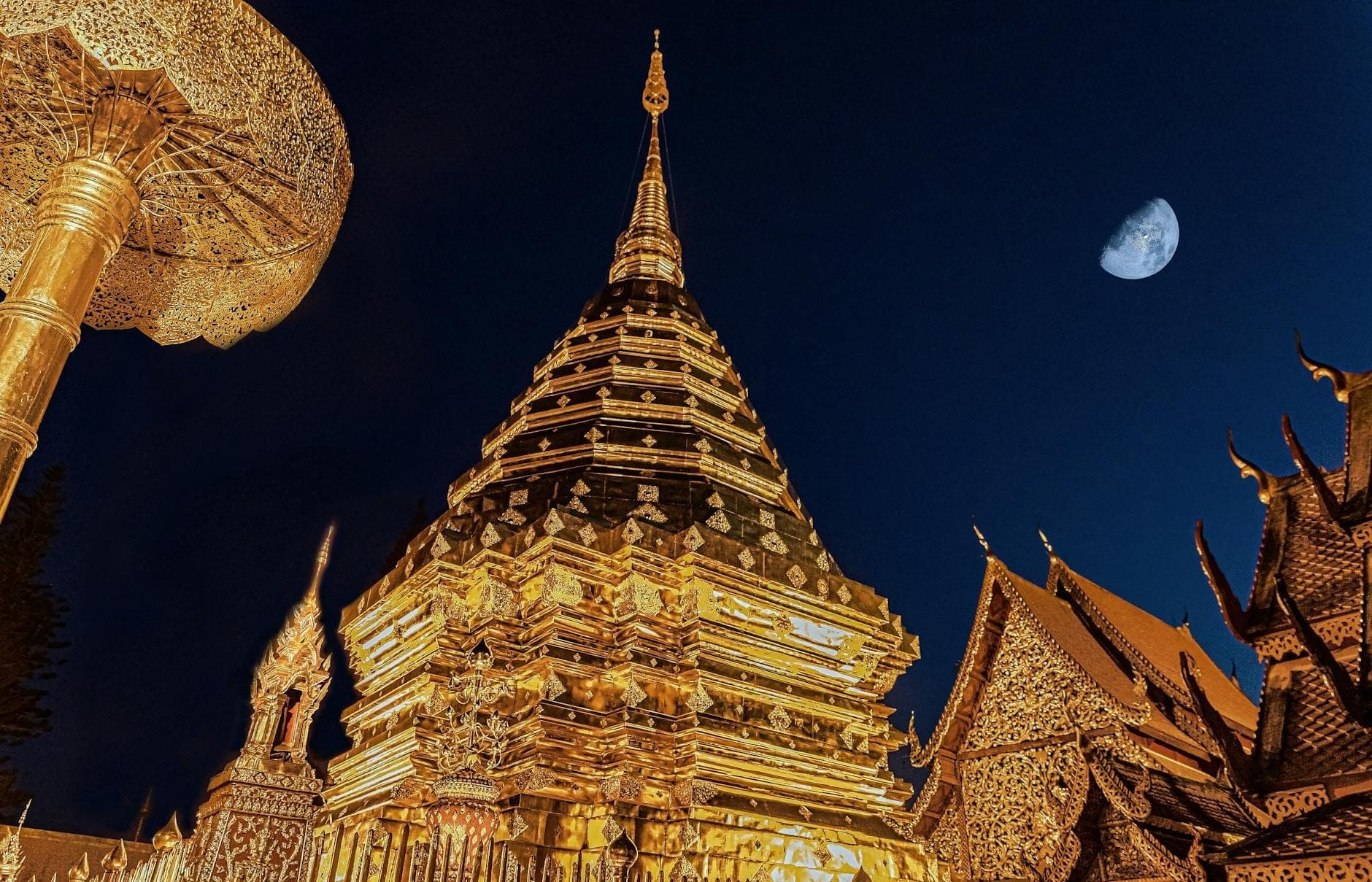 Stupa dorata del Wat Phra That Doi Suthep illuminata di notte con la luna in un cielo blu scuro, che mostra l'esclusiva atmosfera del tempio.