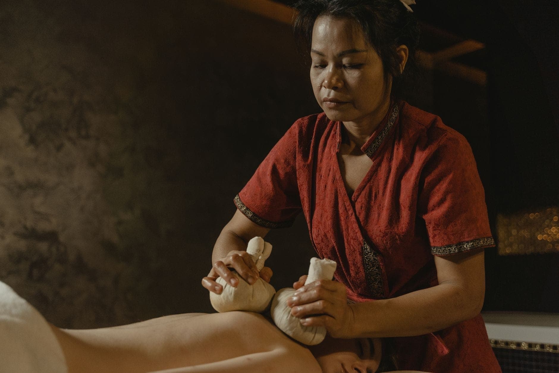 Una donna in abito tradizionale esegue un massaggio con impacchi alle erbe in una spa dall'illuminazione soffusa, evocando le tradizioni del benessere Thai.