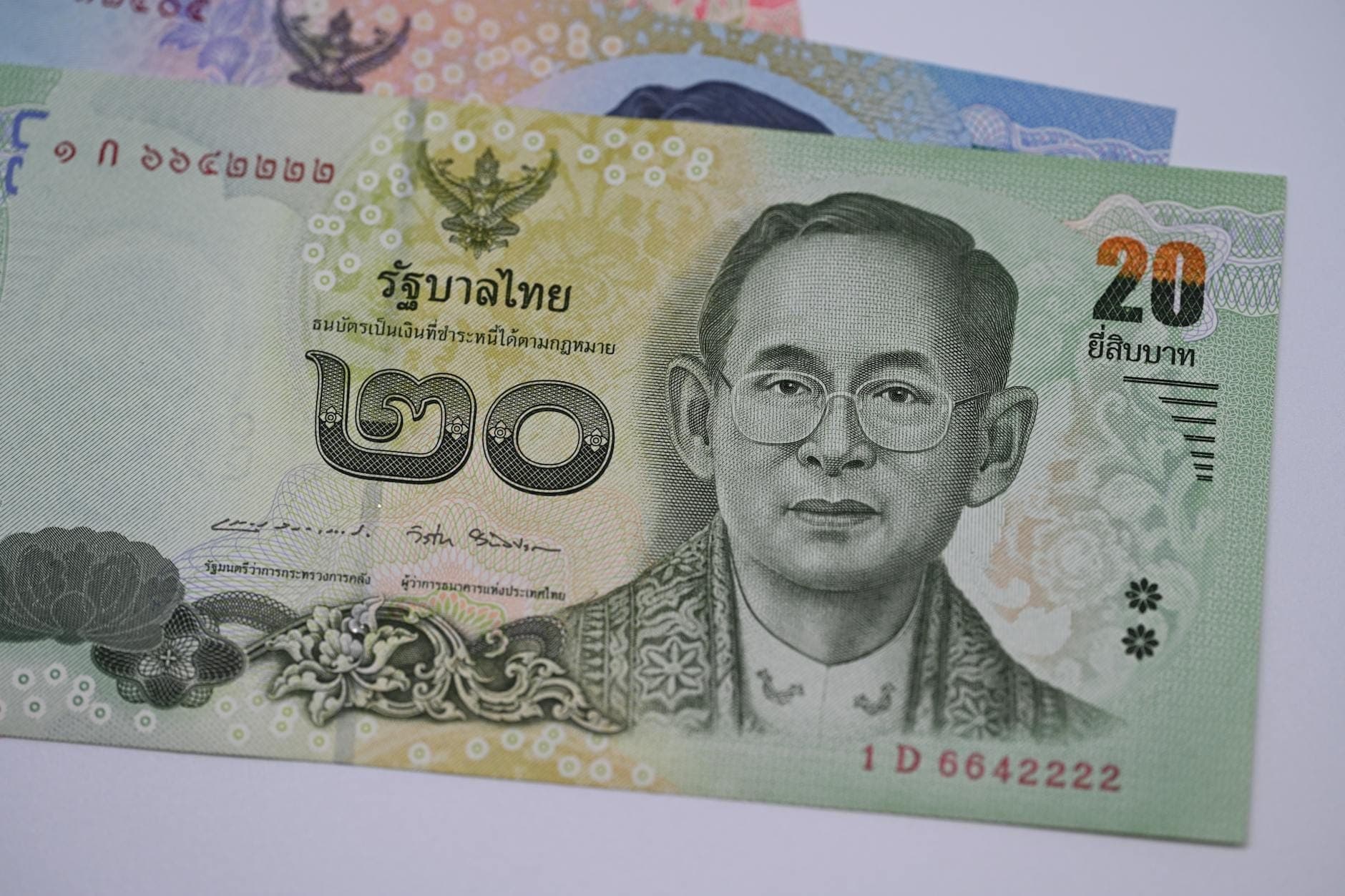 Arka planda hafifçe görünen diğer baht banknotlarıyla birlikte, üzerinde Tayland kralının portresi bulunan 20 Tayland bahtı banknotunun yakın çekimi.