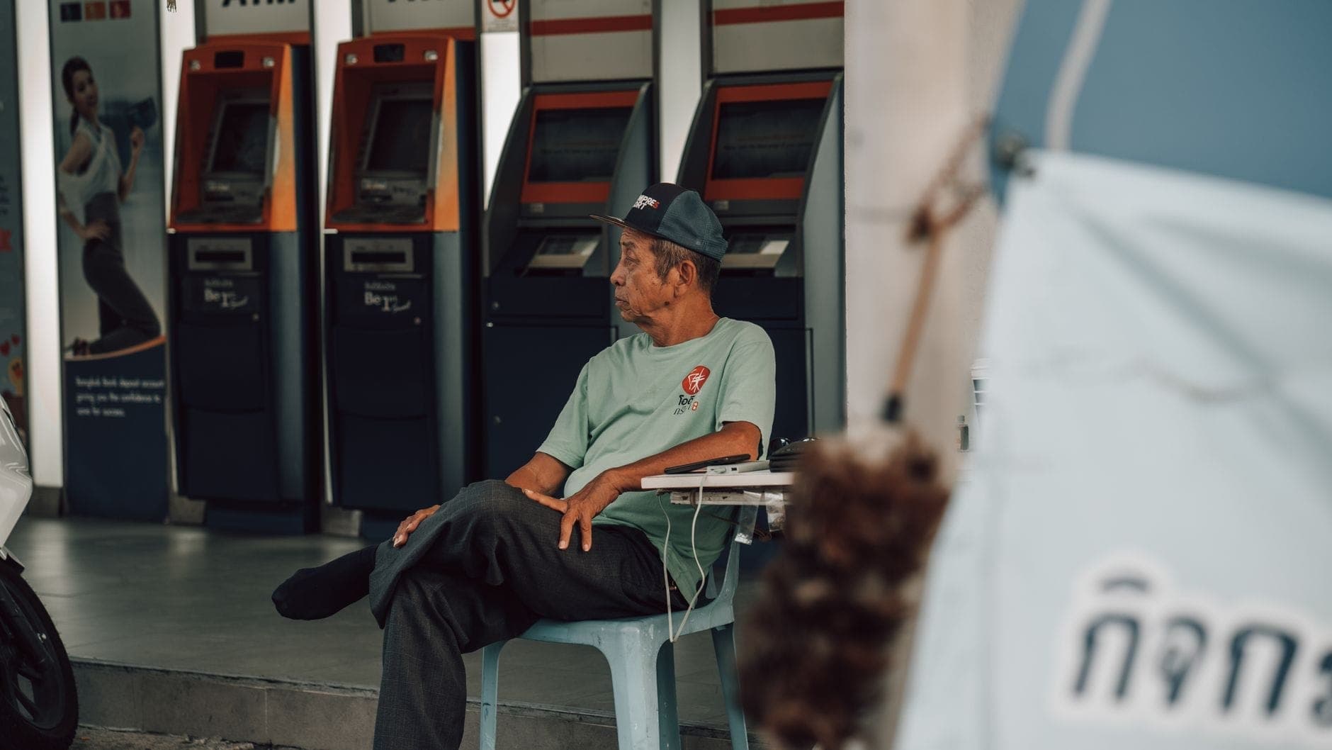 Üzerinde Tayca yazı bulunan modern ATM sırası, yakında oturan bir adam, sahne belirgin biçimde Güneydoğu Asya görünümünde ve kentsel Chiang Mai'ye özgü.