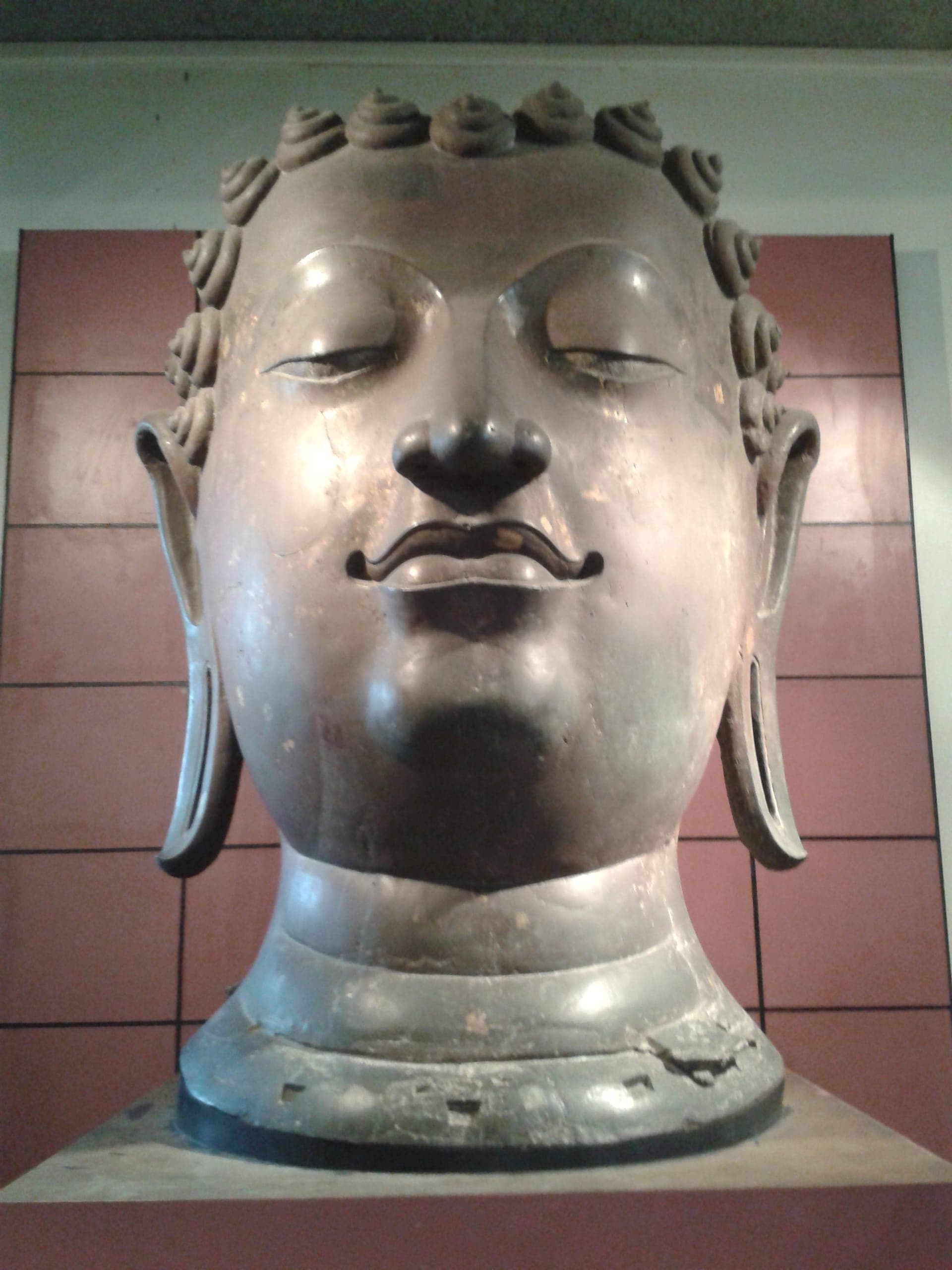 Große Bronzekopfskulptur eines Buddha, ausgestellt vor einer rotgekachelten Wand im Chiang Mai National Museum, unter weicher Innenbeleuchtung.