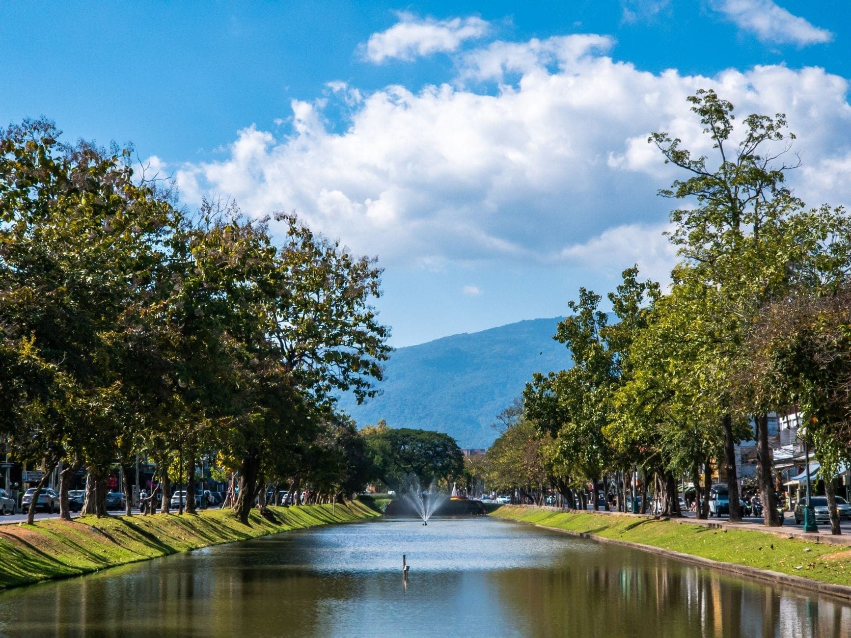 Blick entlang eines baumgesäumten Kanals mit einem Springbrunnen, parkenden Autos und bewaldeten Bergen unter strahlend blauem Himmel in Chiang Mai.