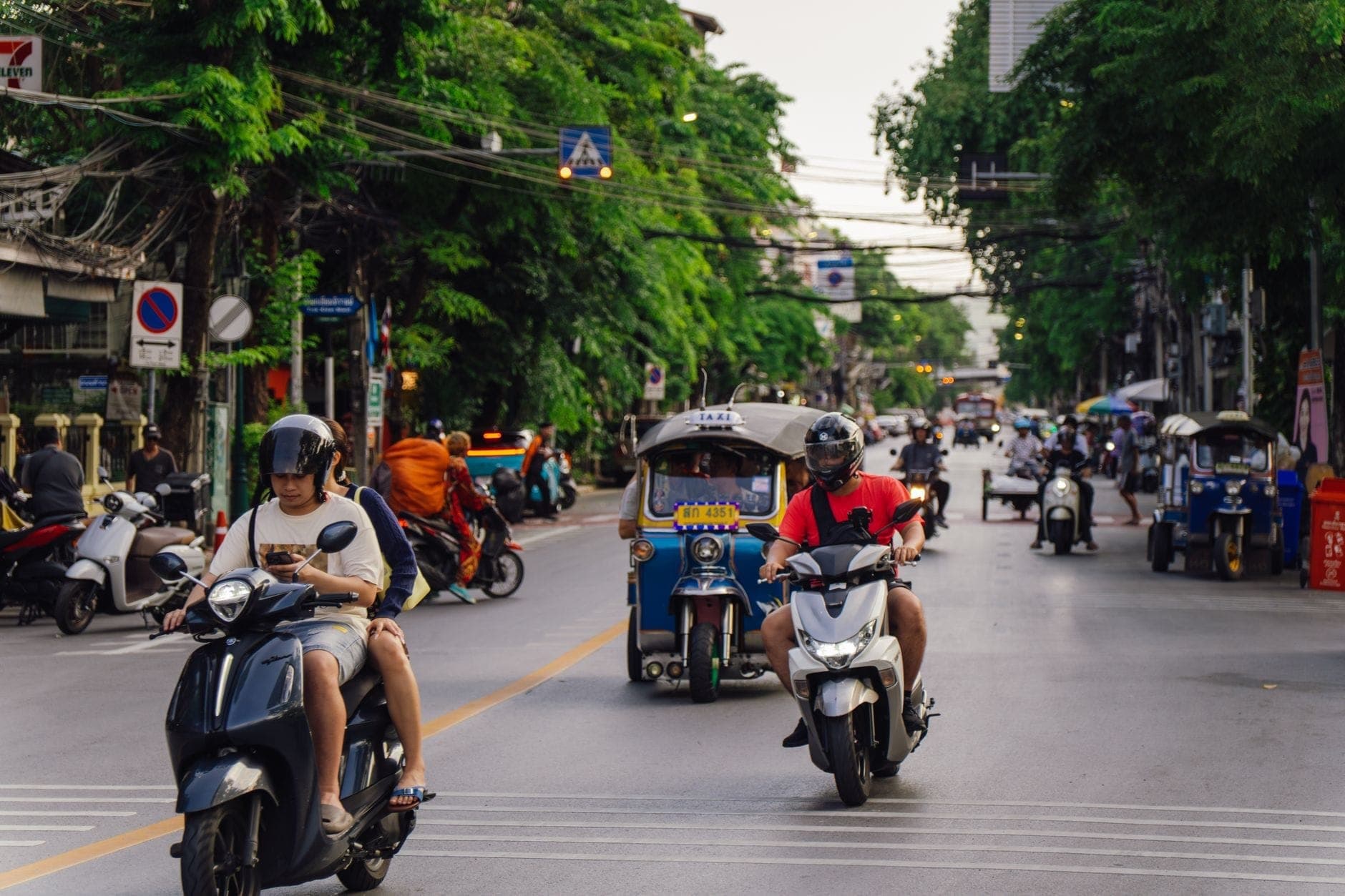 Ruchliwa ulica w Chiang Mai z kilkoma osobami jadącymi na motocyklach, tuk tukiem i miejskimi budynkami w tle.