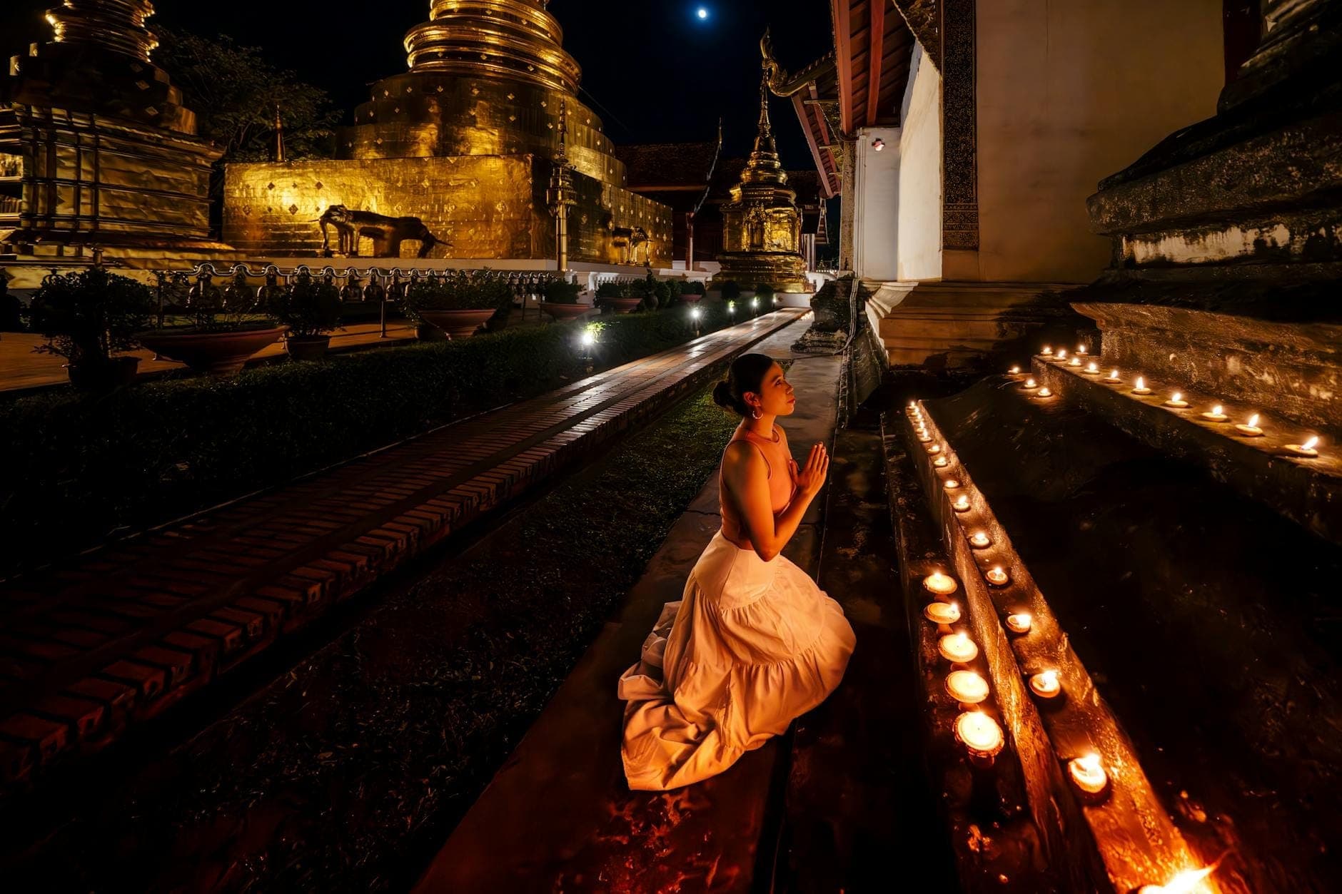 Gece altın bir tapınağın yanında mumlarla dua eden diz çökmüş bir kadın, Chiang Mai'deki Songkran'ın manevi geleneklerini yansıtıyor.