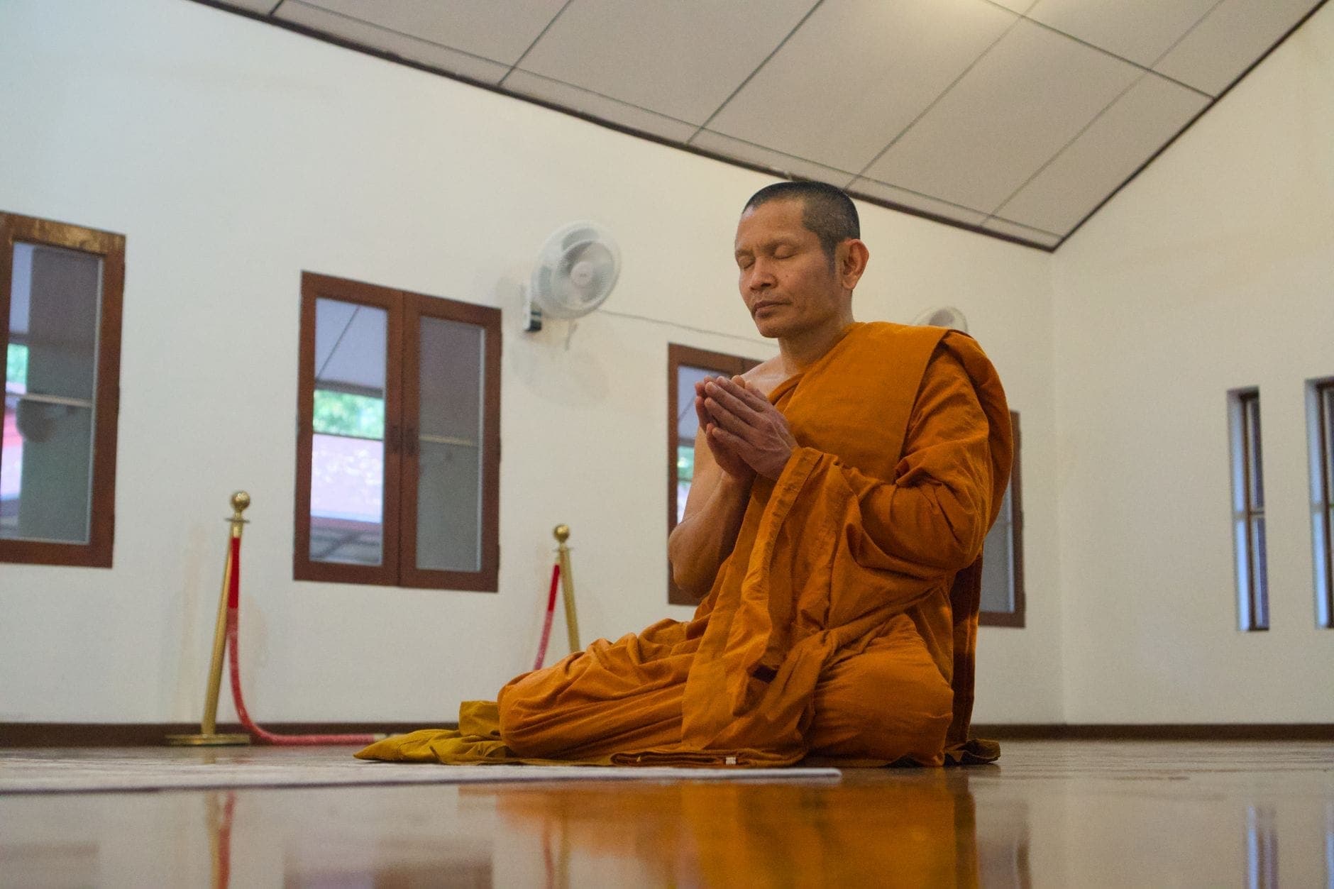 Ein buddhistischer Mönch in orangefarbenen Gewändern meditiert in einem ruhigen Tempelraum mit Holzböden und schlichter Dekoration in Chiang Mai.