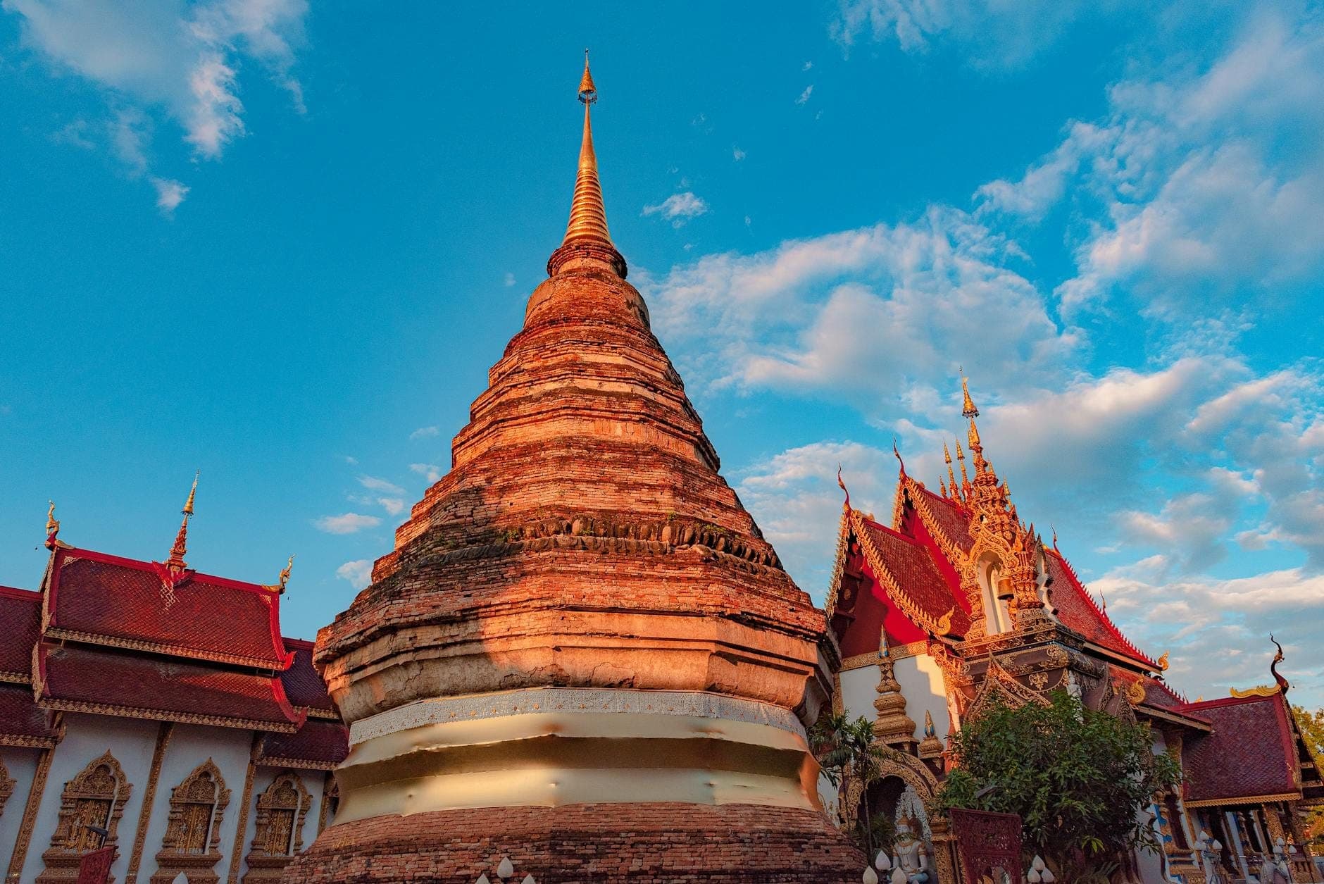 Complejo de templos de Chiang Mai bajo el sol con una estupa dorada y tejados ornamentados bajo un cielo azul con nubes dispersas.