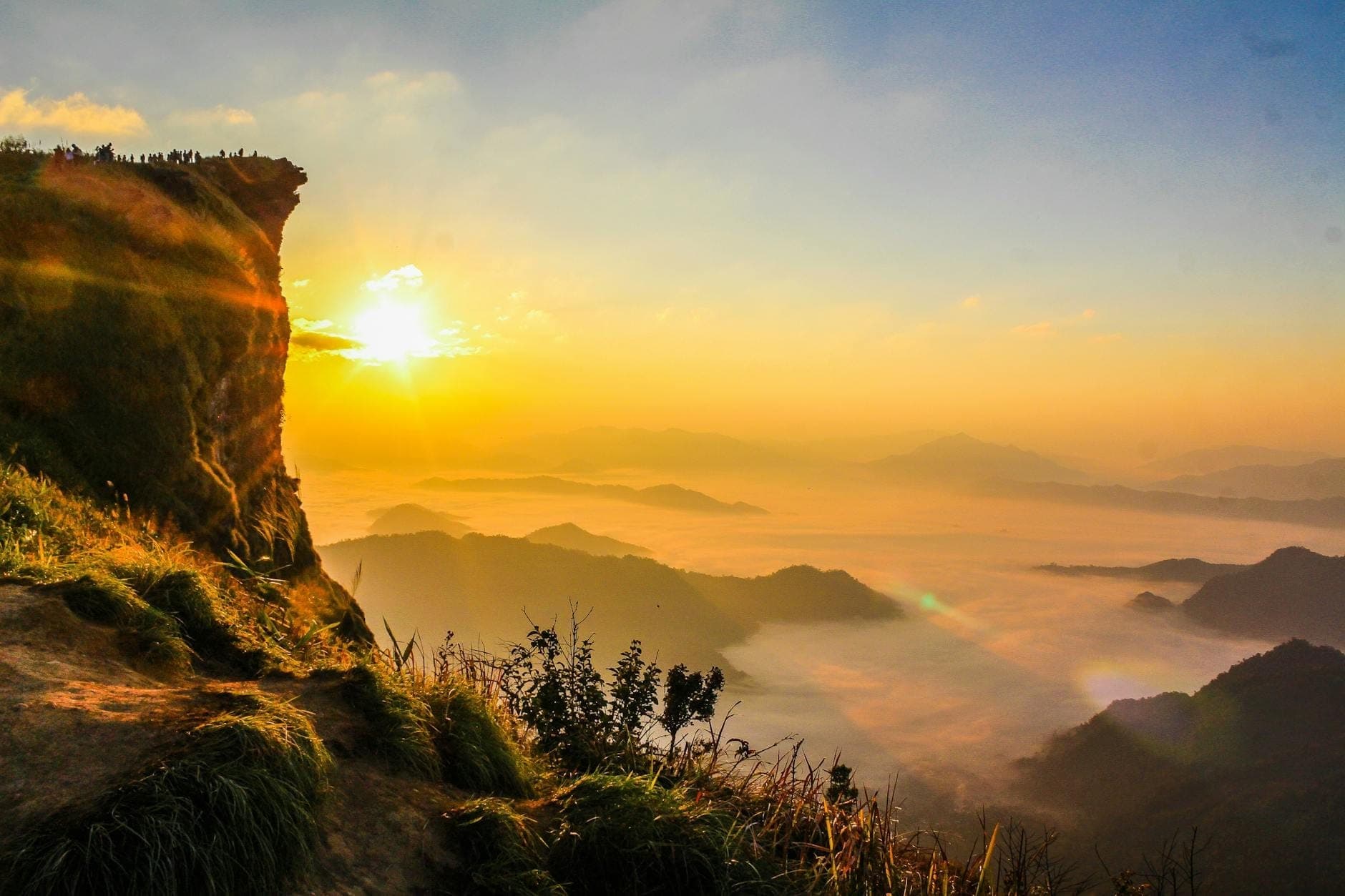 Goldener Sonnenaufgang über nebligen Bergen und Tälern in Nordthailand mit Menschen, die sich auf einer Klippe versammelt haben, um die Szene zu betrachten.