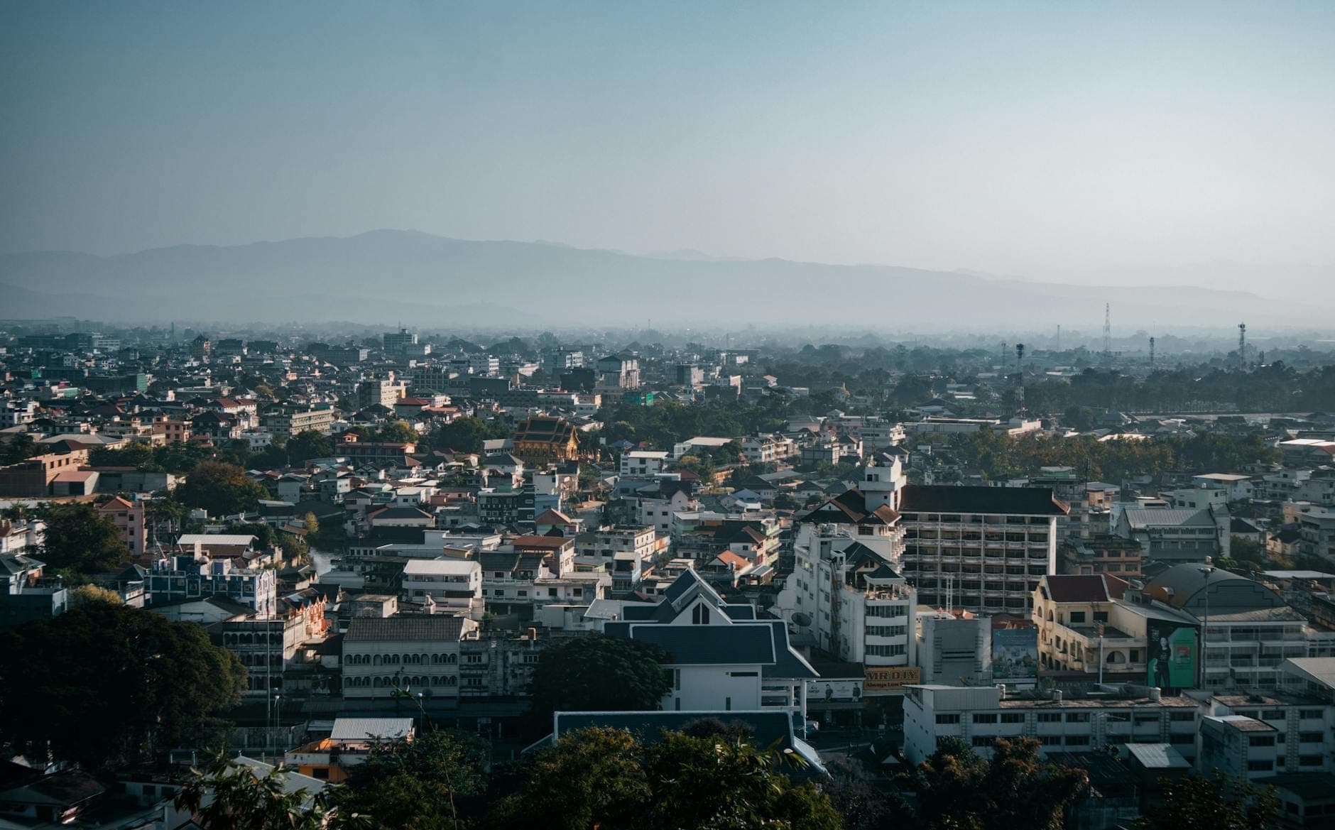 Weitläufige Stadtansicht von Chiang Rai mit dicht gedrängten Gebäuden, einigen höheren Hotels und bewölkten Bergen im Hintergrund.