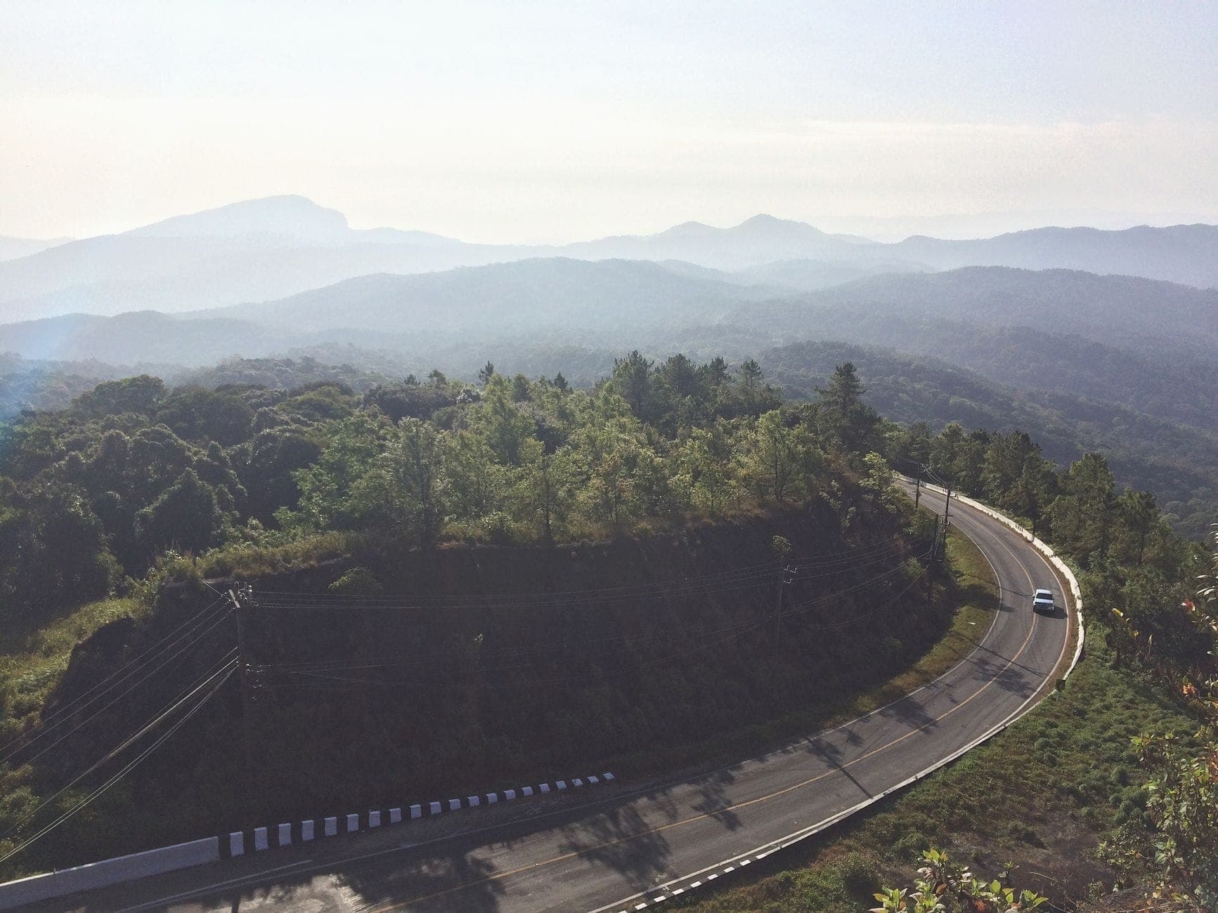 Kurvenreiche Bergstraße mit einem Auto, umgeben von üppigem Grün und nebligen Hügeln auf der Strecke zwischen Chiang Mai und Chiang Rai bei Sonnenaufgang.