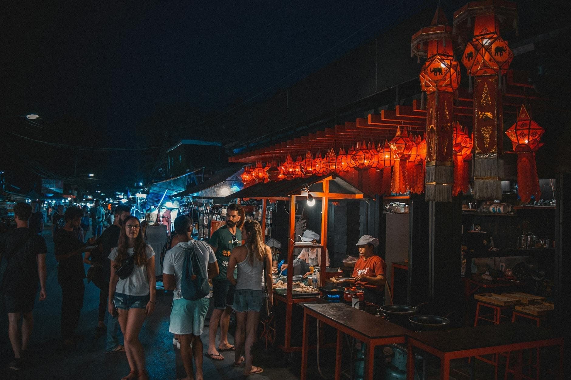 Escena de mercado nocturno con personas caminando junto a puestos y linternas rojas brillantes en lo alto, capturando una animada calle en Pai, Tailandia.