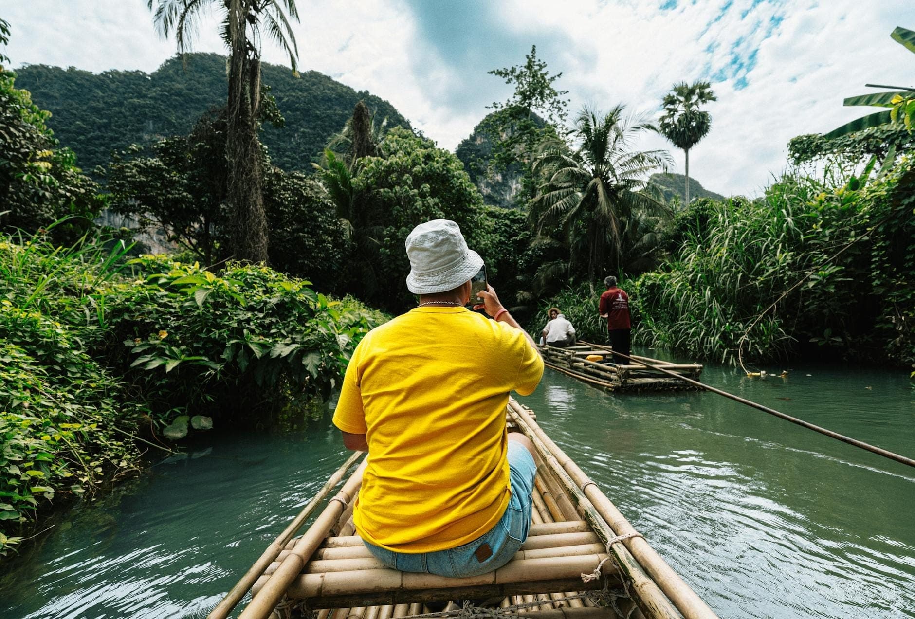 Turisti su una zattera di bambù che navigano un rigoglioso fiume nella giungla con una guida locale, circondati da vegetazione verde e montagne lontane.