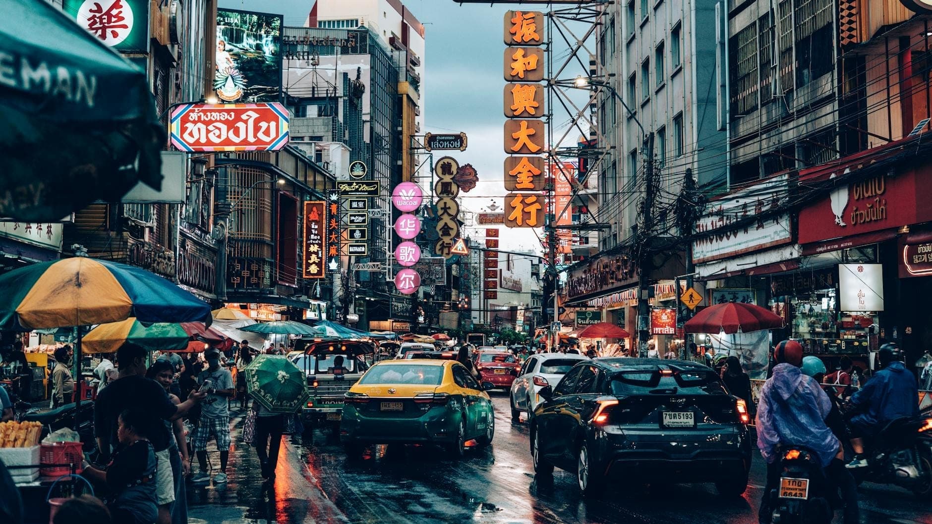 Belebte Straßenszene in Bangkoks Chinatown in der Abenddämmerung mit Neonreklamen, Taxis, Motorrädern, Straßenhändlern und Menschen unter bunten Regenschirmen.