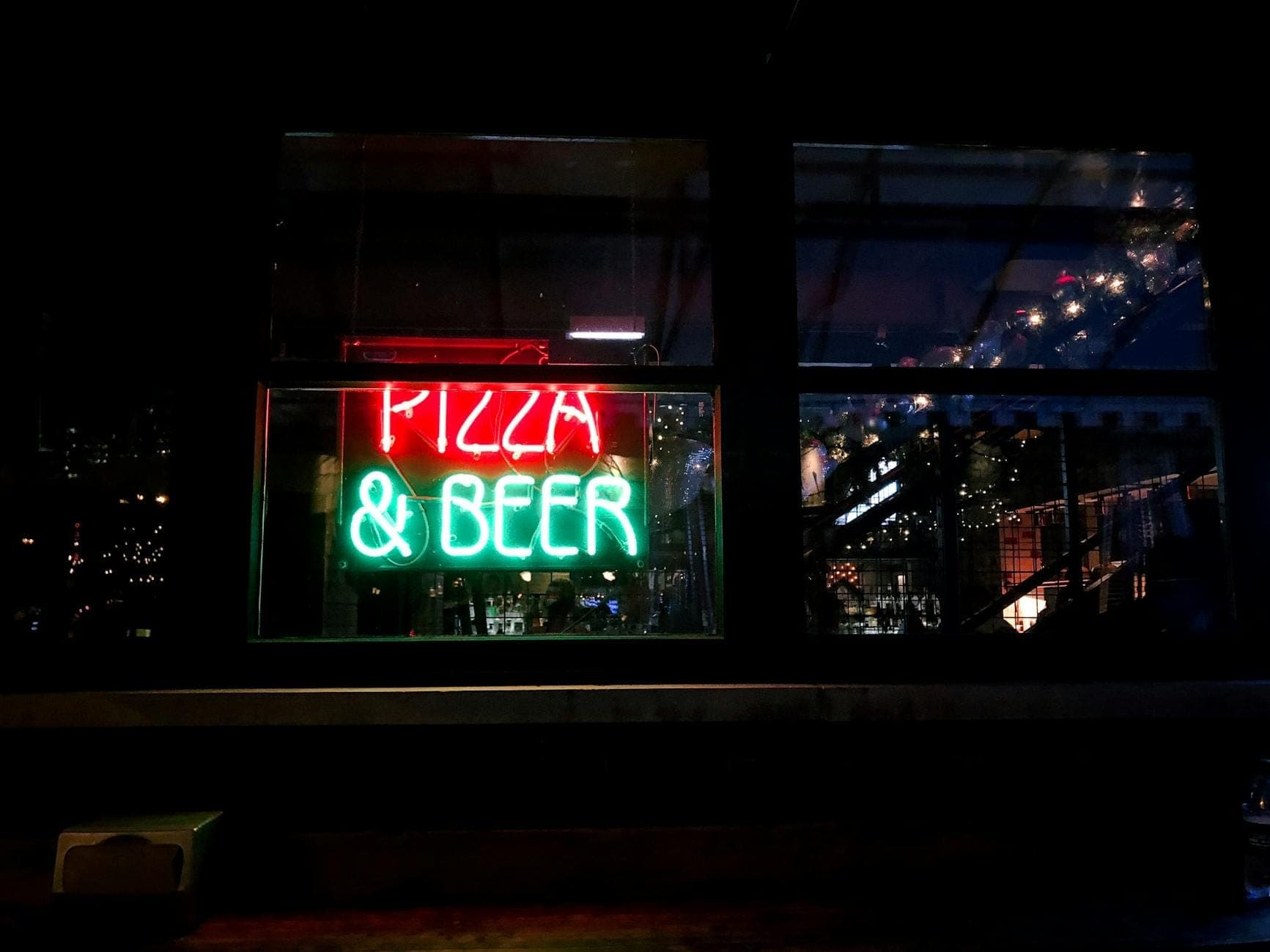 Neon sign in a window at night reading Pizza & Beer with reflections of city lights, suggesting a lively evening urban scene.
