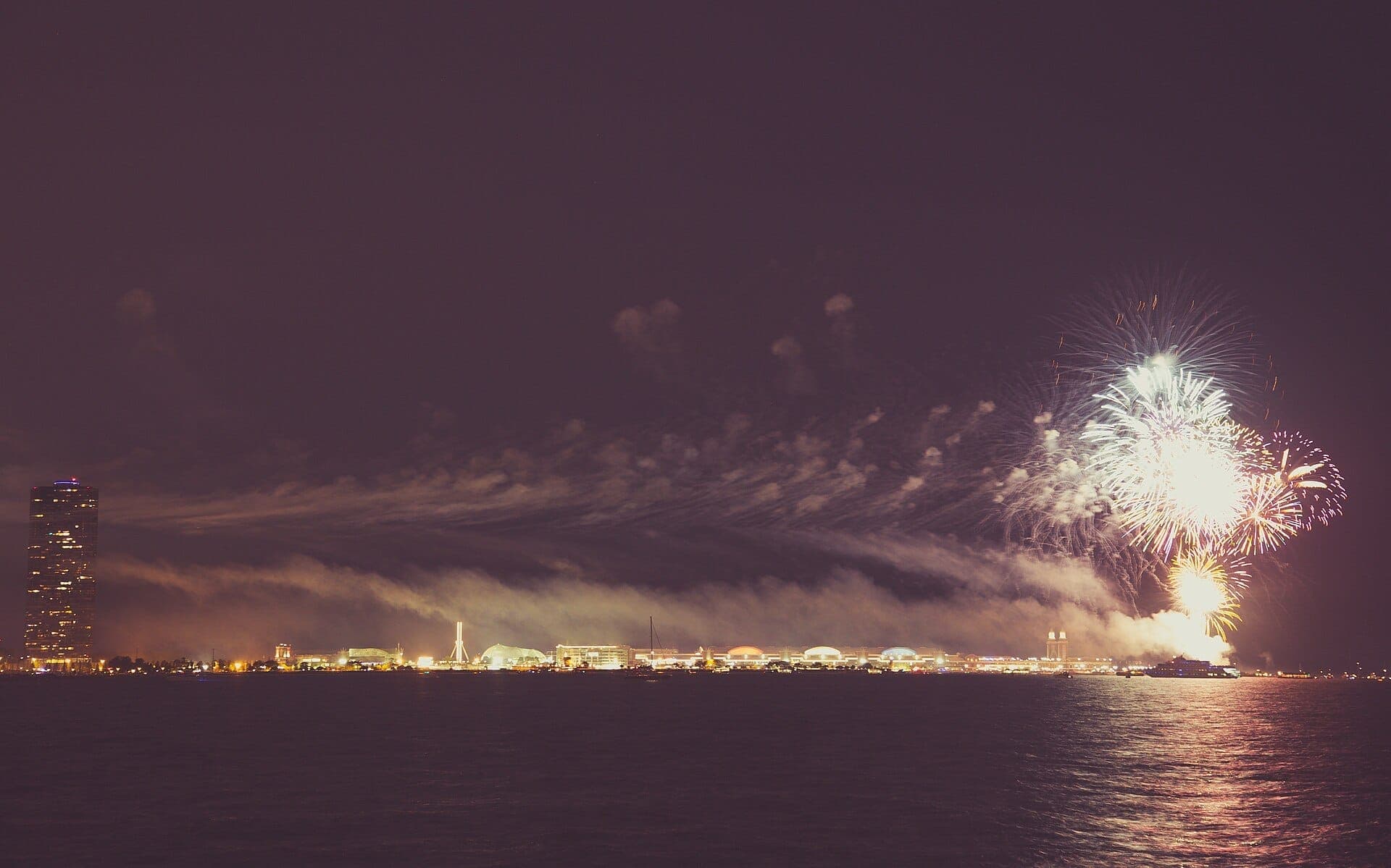Colorful fireworks explode over Navy Pier and Lake Michigan at night, with a high-rise and city lights visible along the shoreline.