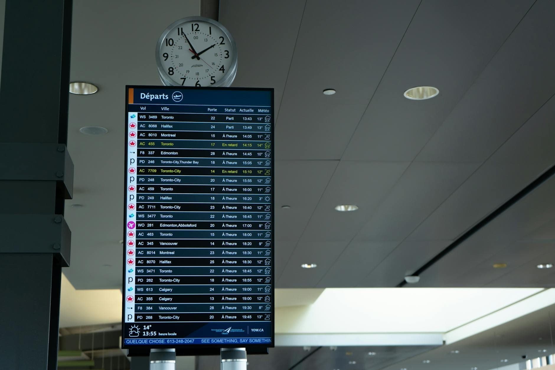 Abflugtafel am Flughafen mit mehreren Airline-Flügen und Reisezielen, mit einer Uhr darüber und der modernen Terminaldecke im Hintergrund.