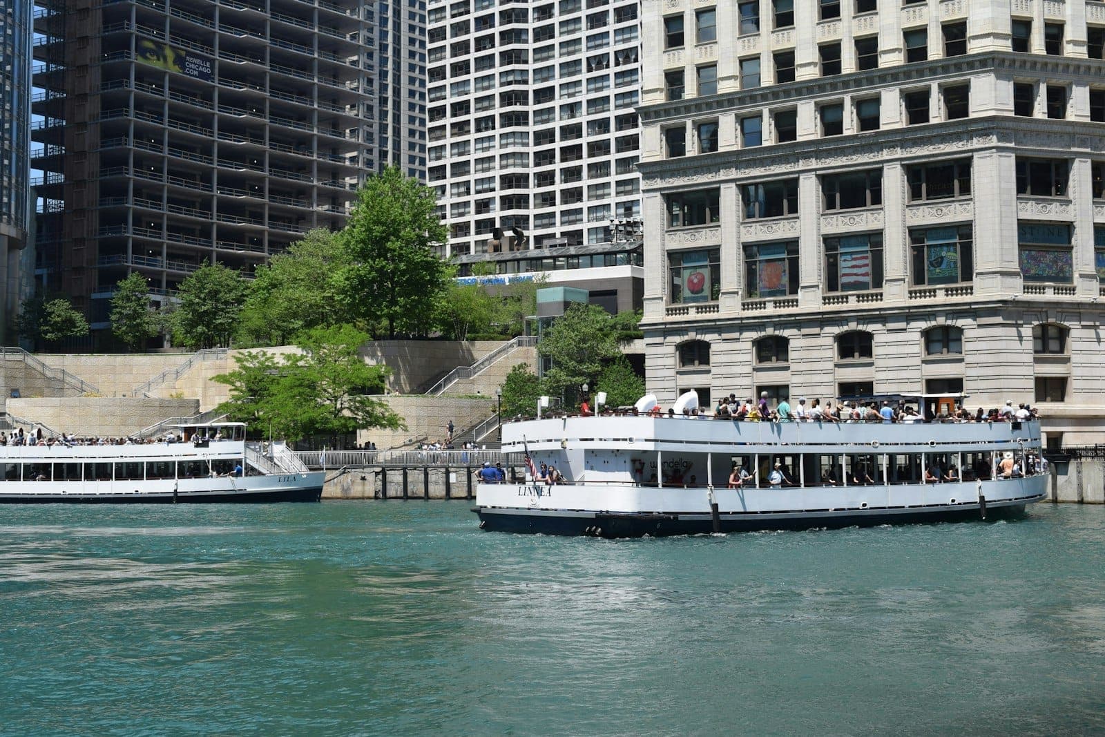 Dos barcos de recorrido arquitectónico atracados junto al Chicago Riverwalk, con edificios de la ciudad al fondo en un día soleado.