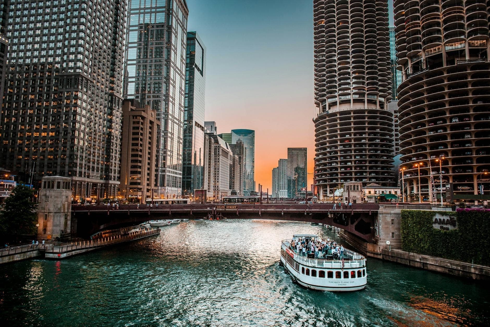 A Chicago river cruise boat moves up the river at sunset, surrounded by iconic downtown skyscrapers and city bridges under a vibrant sky.