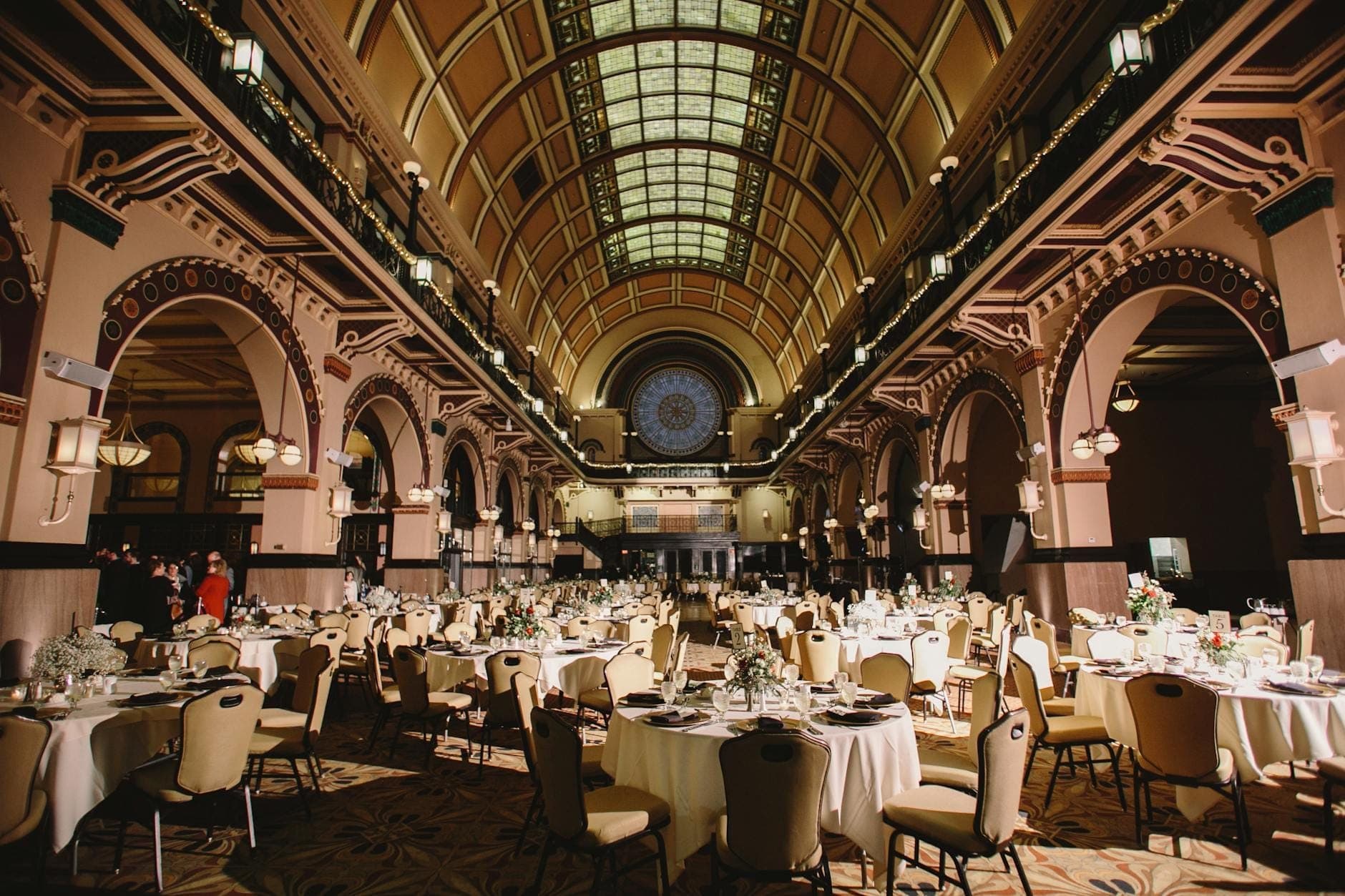 Elegant historic hall with decorative arches, round tables set for an event, warm lighting, and a vaulted glass ceiling.