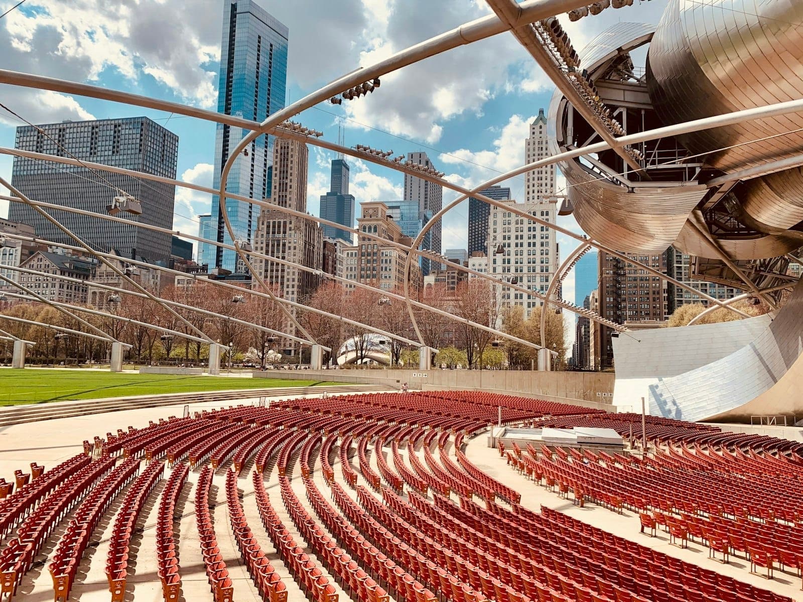 Empty outdoor seating area of Jay Pritzker Pavilion in Millennium Park with Chicago skyline in background, under arched steel trellis.