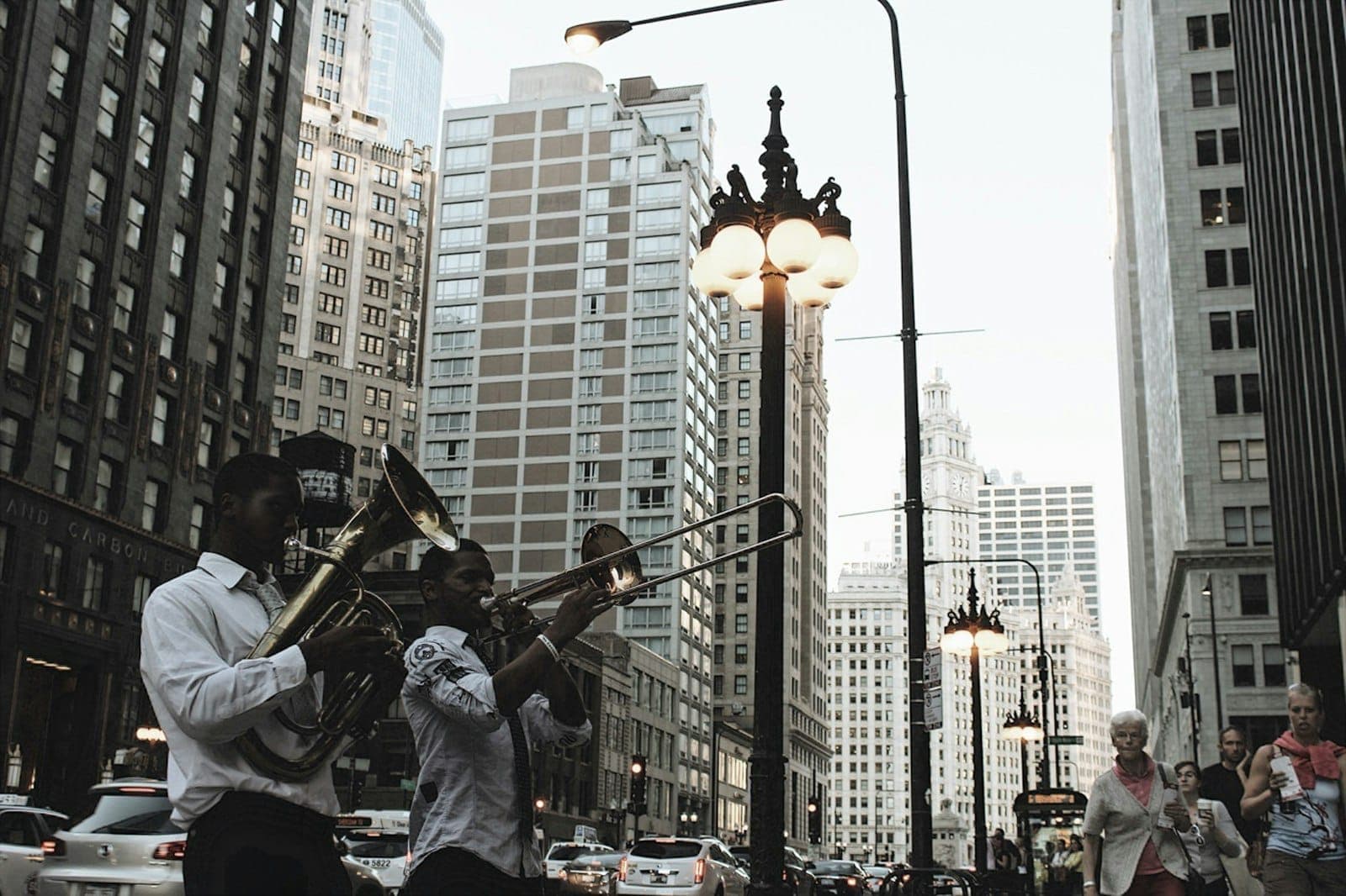 Street musicians play brass instruments in downtown Chicago, surrounded by tall buildings, glowing street lamps, and city traffic in the background.