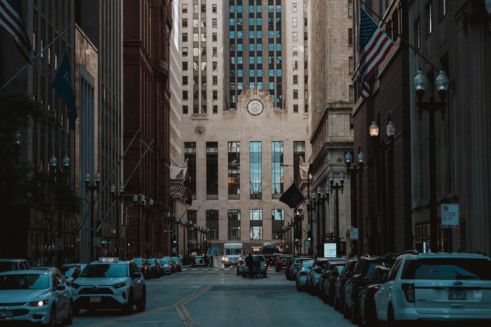 Street-level view of the Chicago Board of Trade Building framed by skyscrapers and American flags, with cars lining LaSalle Street in downtown Chicago.