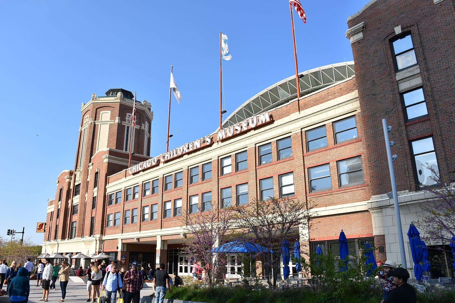 The exterior of the Chicago Children's Museum at Navy Pier, with red brick facade, flags, and families gathered outside on a sunny day.