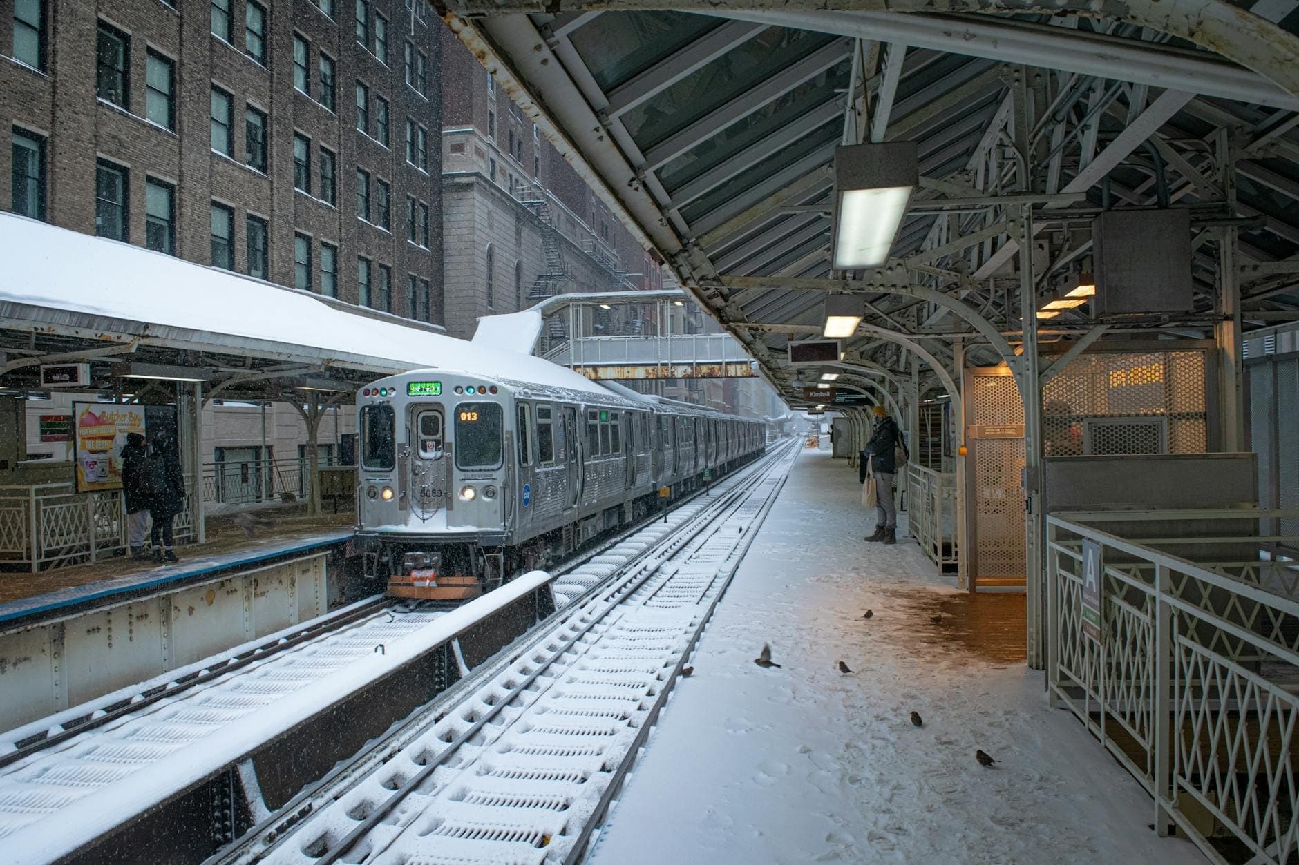 Chicago CTA train arriving at a snowy elevated platform in winter with people waiting, surrounded by urban buildings.