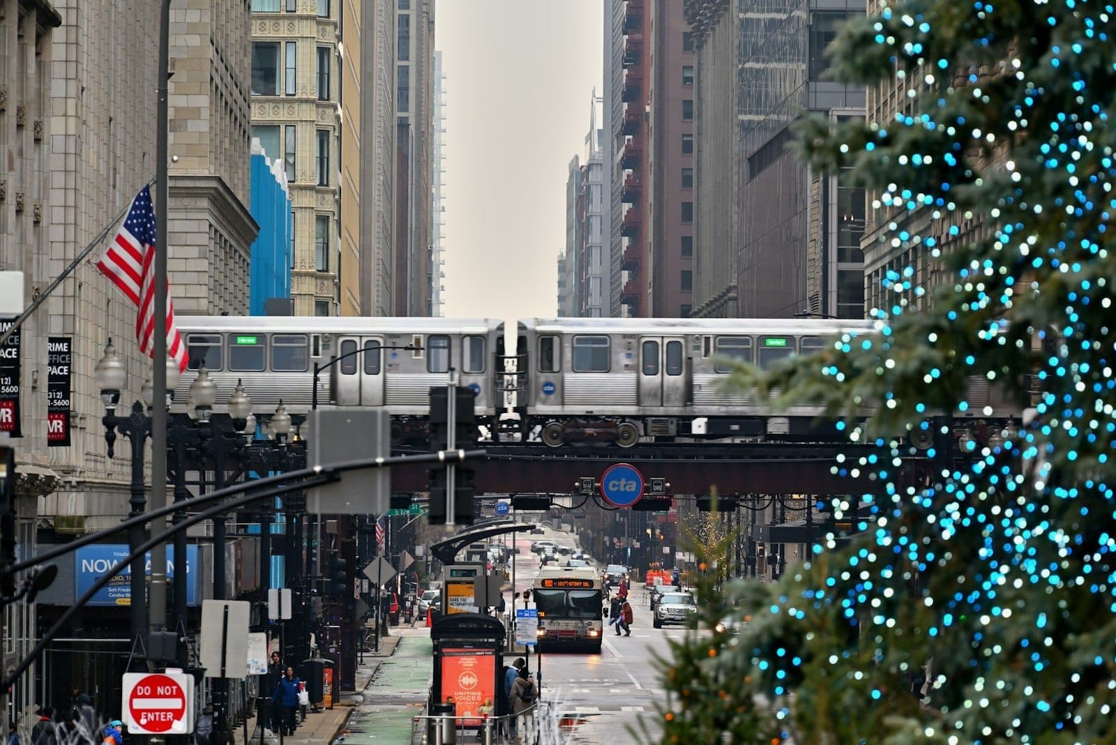 View of downtown Chicago street with Christmas tree decorated with blue lights, elevated train passing above, and tall buildings on a cloudy winter day.