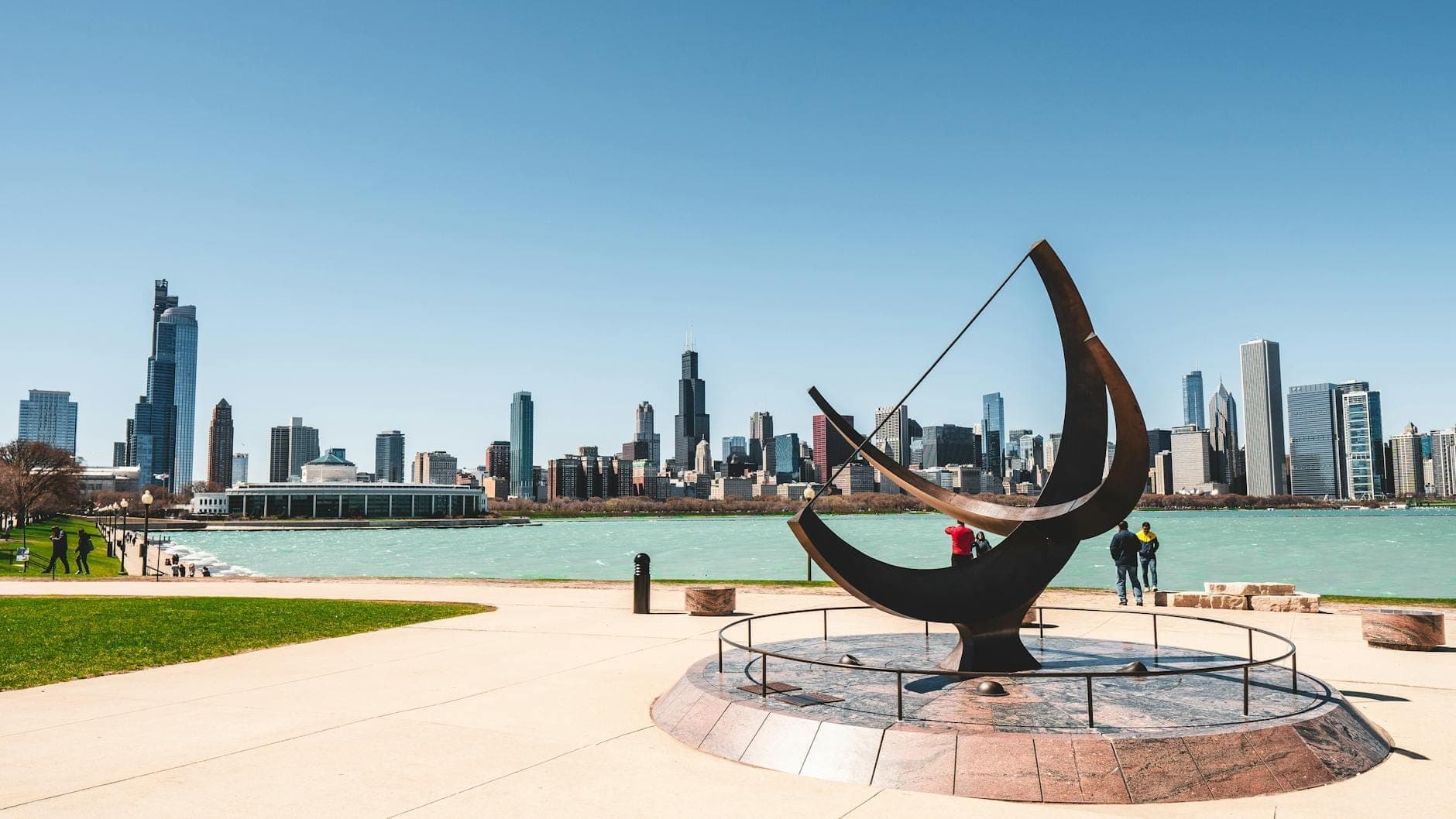 Wide view of Chicago skyline from the shore, with the lake in the foreground, a sundial sculpture nearby, and people enjoying the city scenery.
