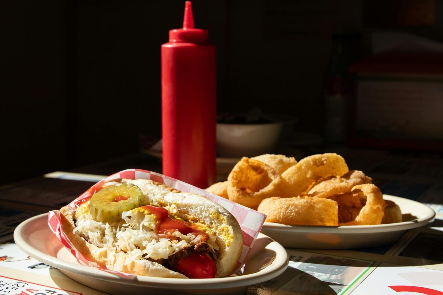 A classic Chicago-style hot dog with pickles, tomatoes, onions, and mustard in a bun, served with onion rings and a red condiment bottle.