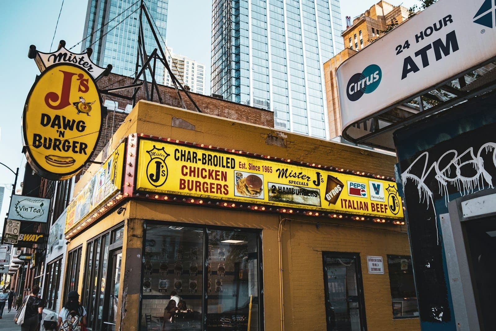 A colorful Chicago street food stand with signs for classic dishes like Italian beef and burgers, set against a backdrop of city buildings and skyscrapers.