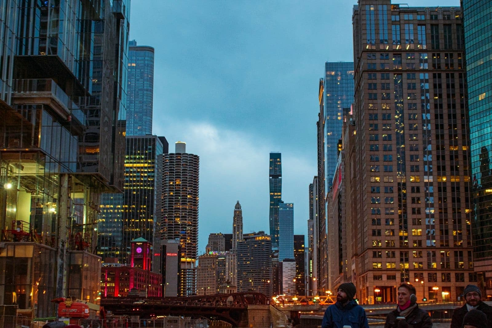 Downtown Chicago cityscape at dusk with lit skyscrapers, river, and people walking on a lively urban street.