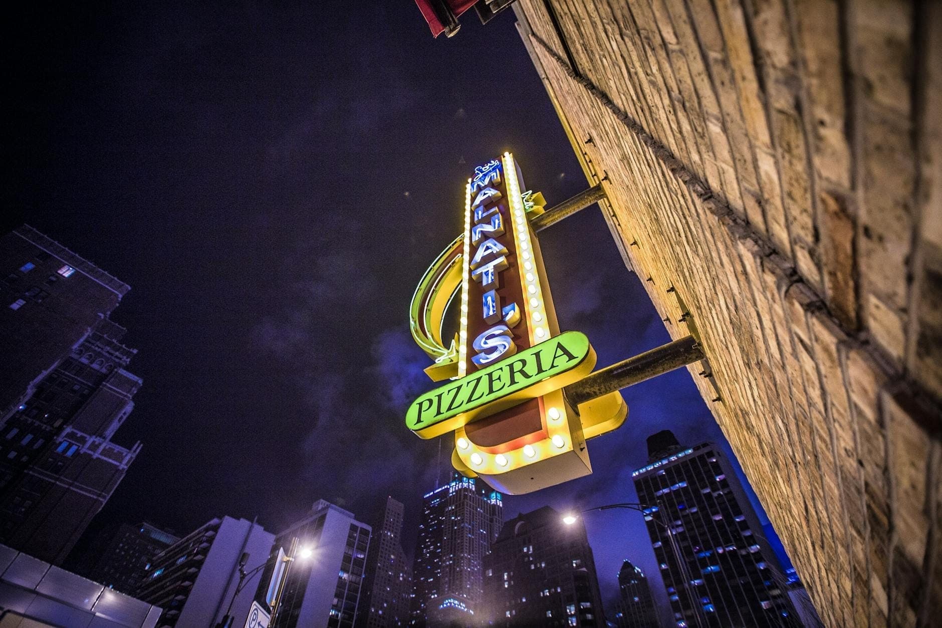 Night view of a classic Chicago pizzeria sign with downtown skyscrapers in the background, depicting the city’s dining scene.