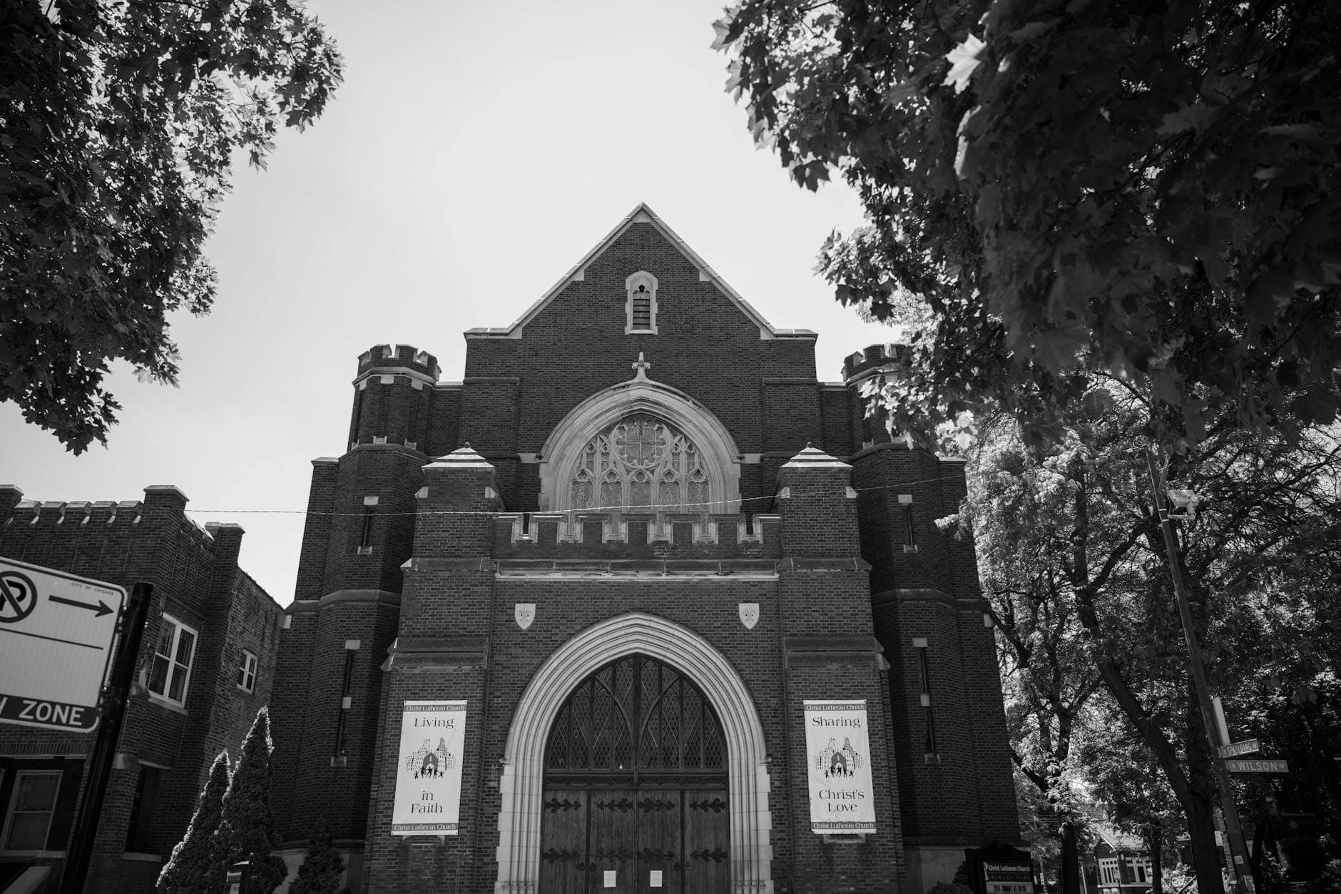 A historic brick church facade with arched doorway and banners, framed by leafy trees, in black and white.