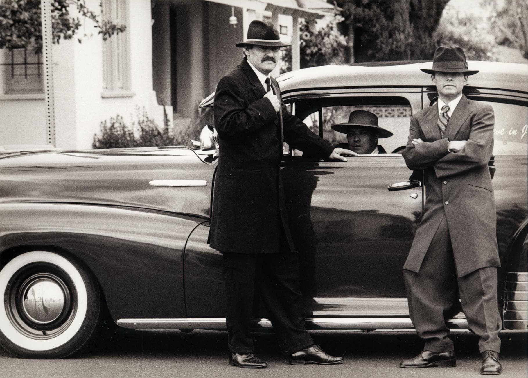 Three men in vintage suits and fedoras stand by a classic 1930s car outside a Chicago-style building, black and white photo.