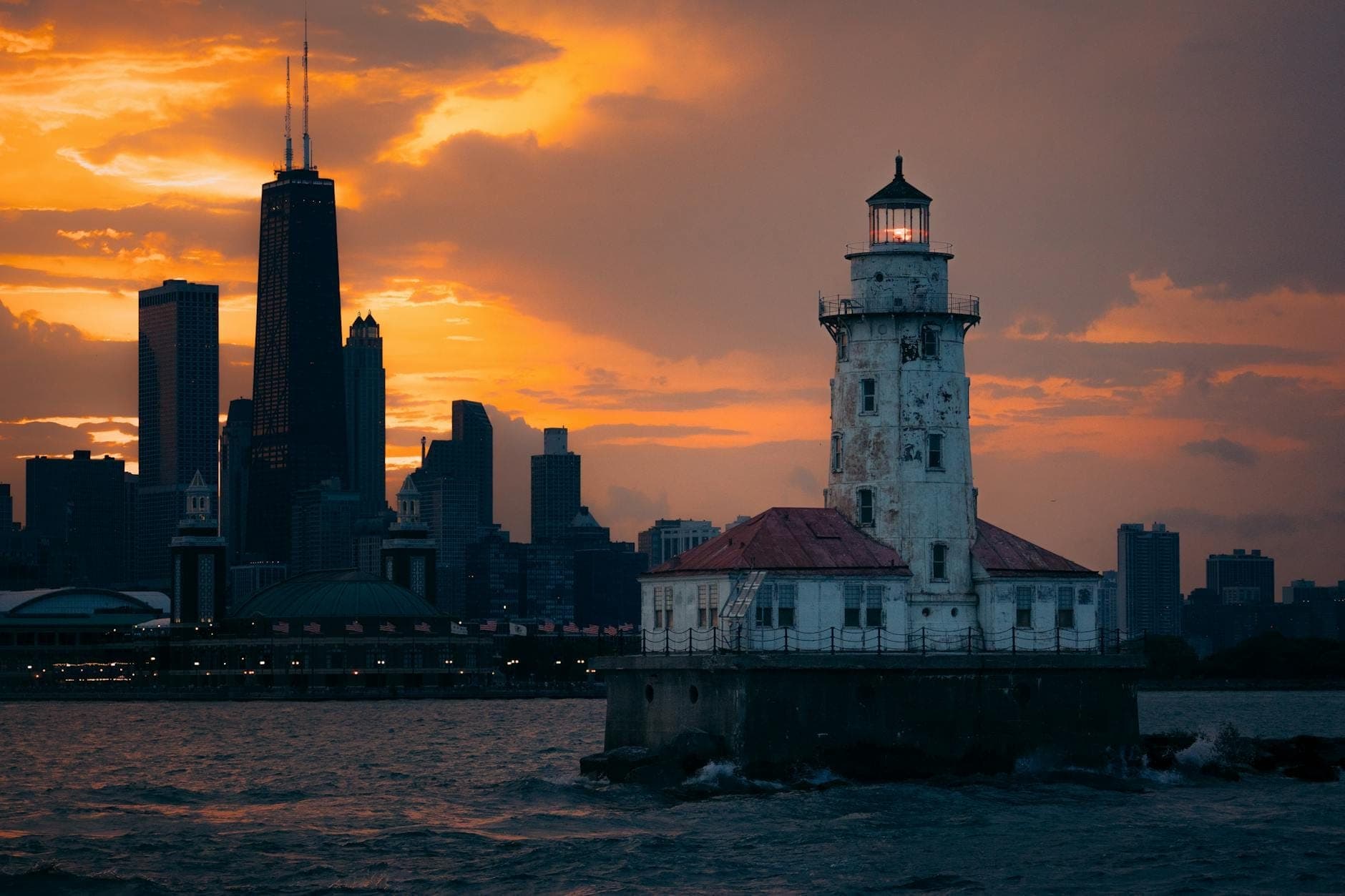 Chicago Harbor Lighthouse illuminated at sunset with the Chicago skyline and dramatic orange sky in the background, viewed from the water.
