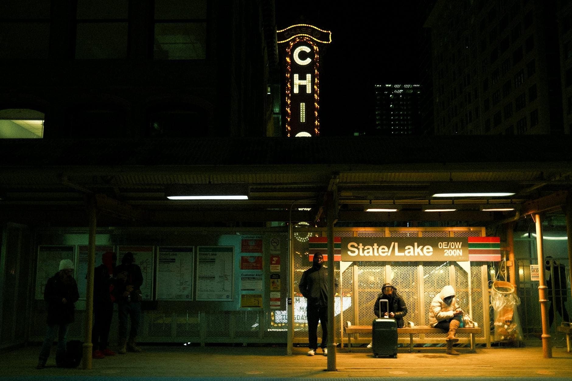Nighttime scene of Chicago's State/Lake elevated train station with people waiting, illuminated signage, and a classic Chicago theater sign in the background.