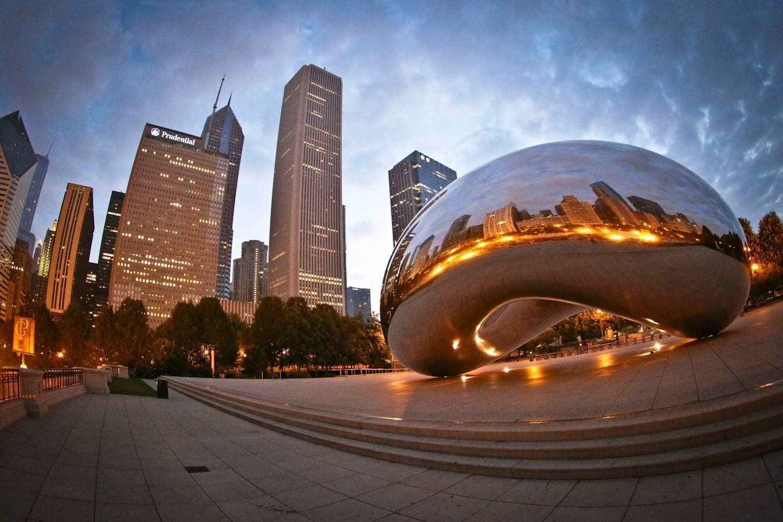 Iconic Chicago skyline at dusk with the Cloud Gate sculpture reflecting the city’s lights in Millennium Park, under dramatic skies.